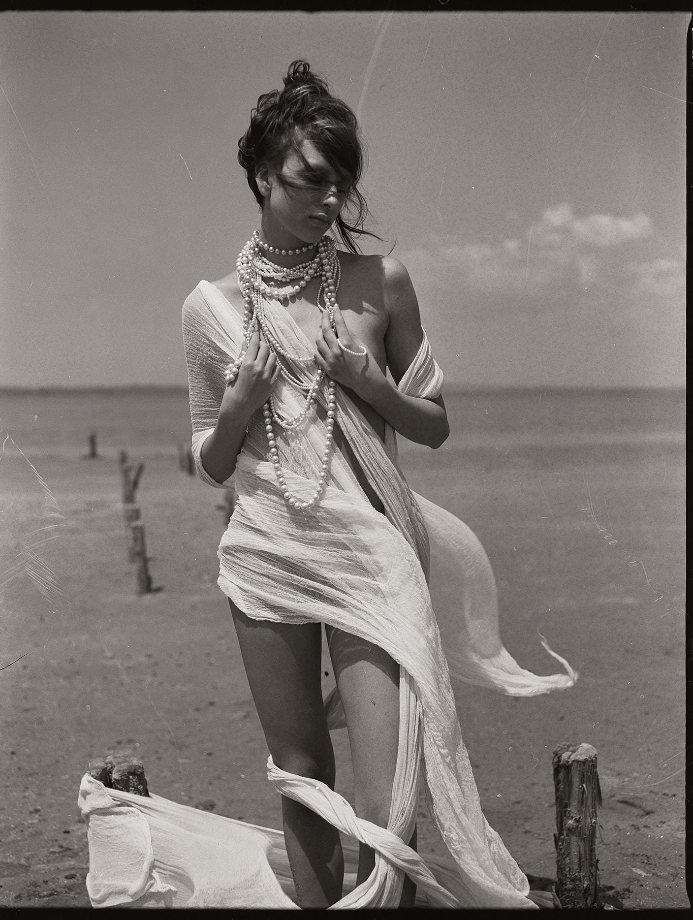 Monochrome portrait of woman in flowing white fabric and layered pearl necklaces on beach.