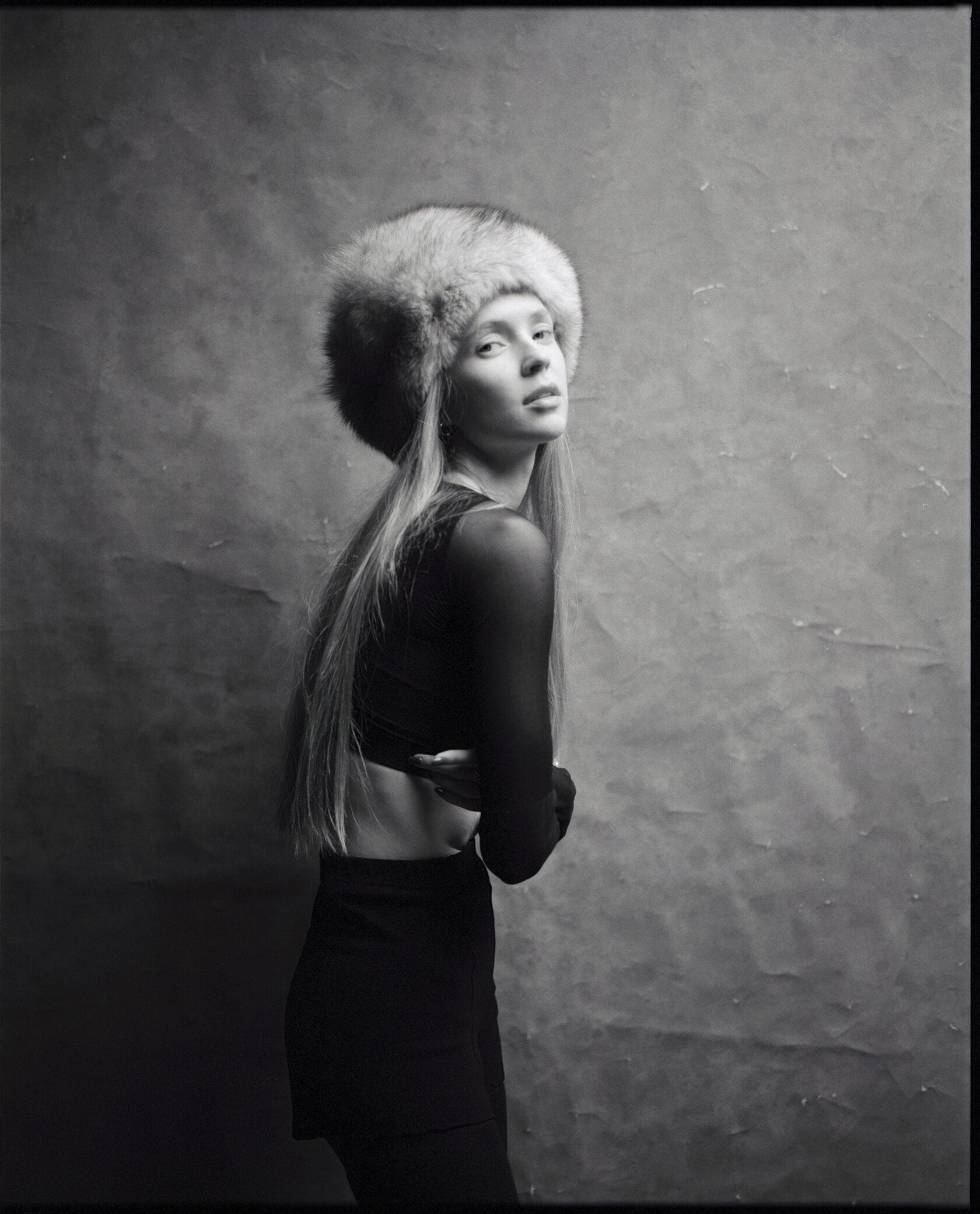 Woman in fur hat and crop top looks over shoulder against mottled studio background.