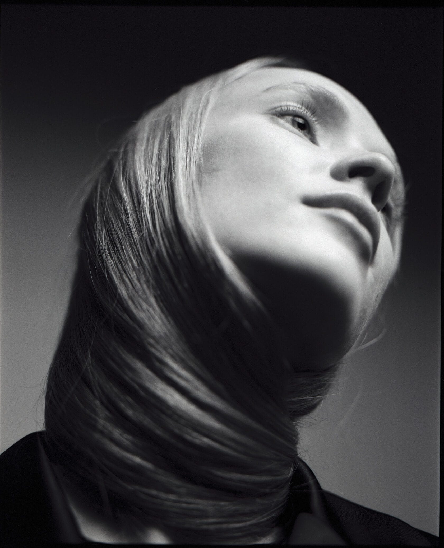 Low angle portrait of woman's face tilted back with hair in motion against dark background.