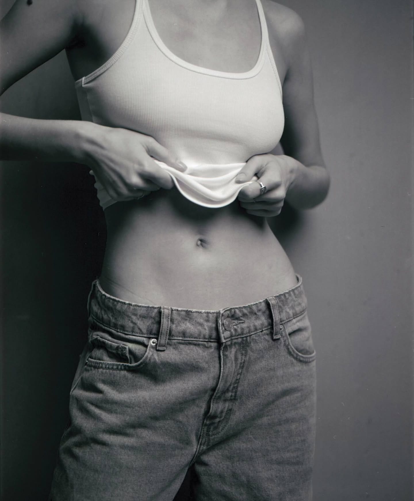 Woman in white tank top pulls hem up showing midriff against gray backdrop.