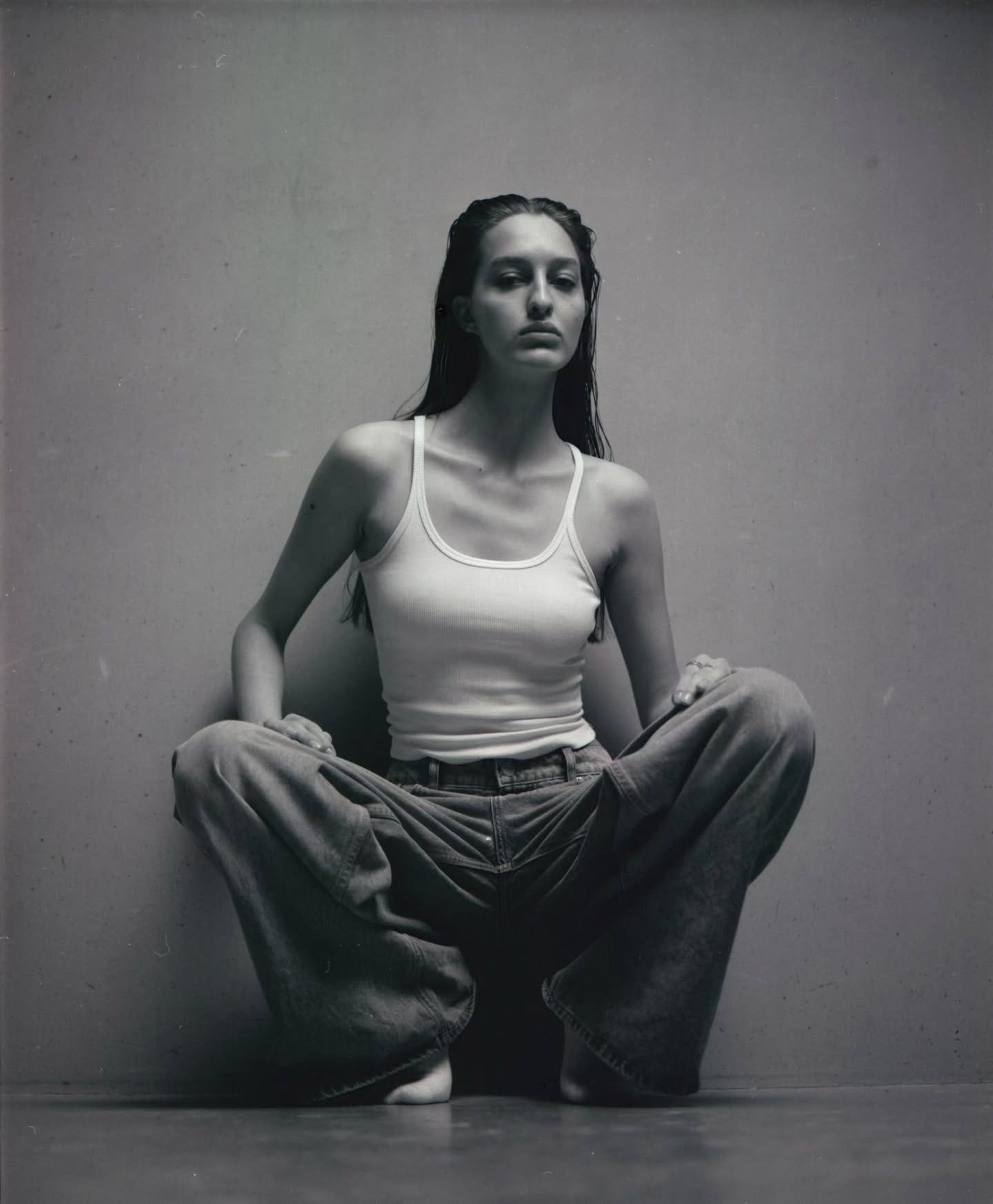 Woman in white tank top sits cross-legged on floor against gray backdrop.