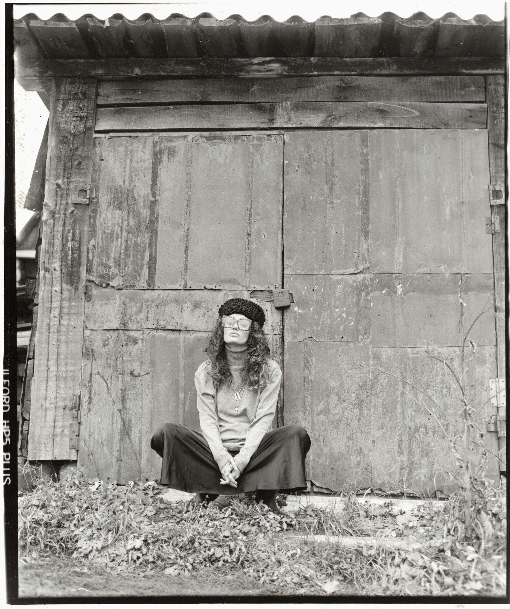 Woman in beret and glasses sits cross-legged against weathered door with film rebate visible.