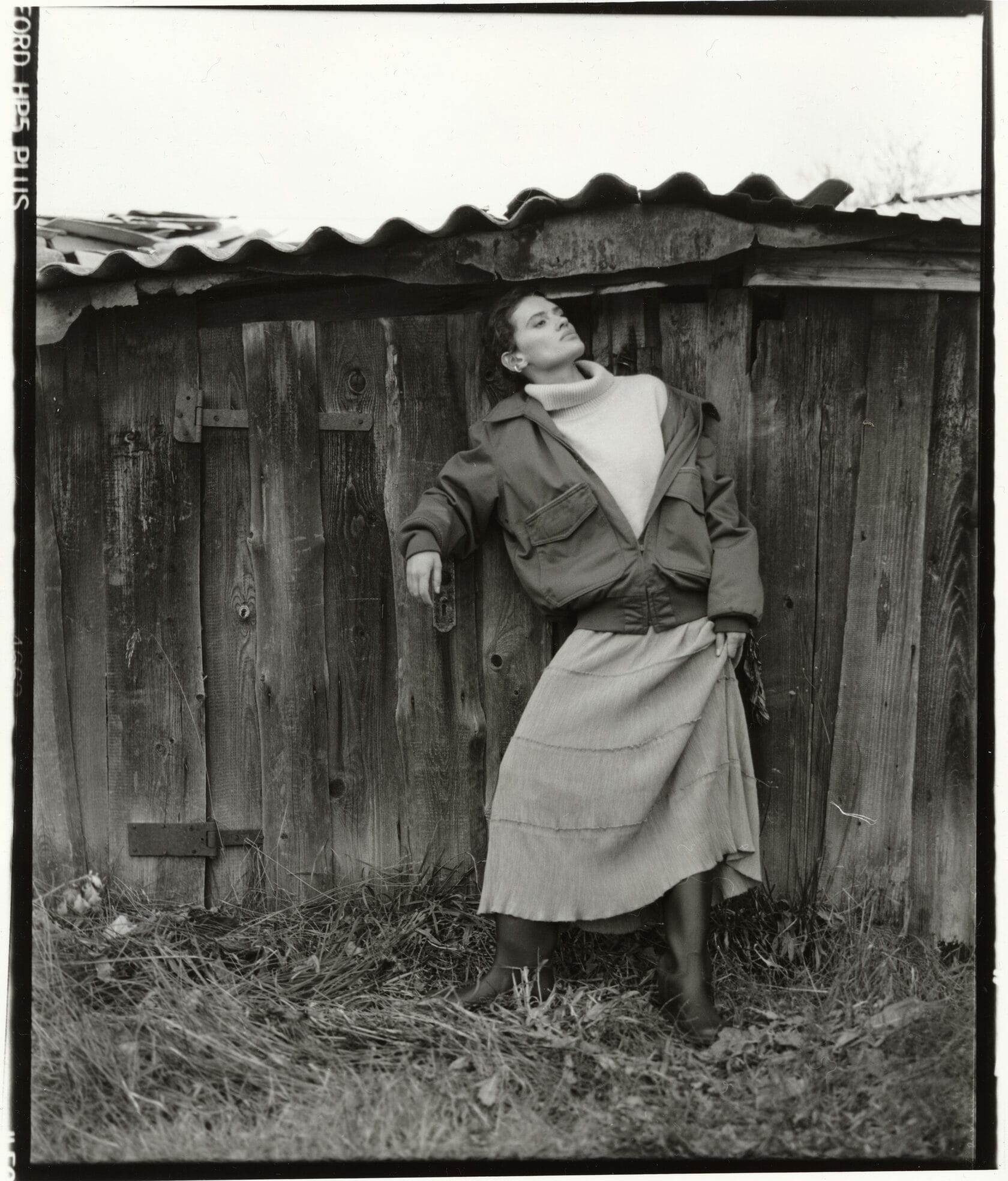 Woman in oversized jacket leans against wooden fence with corrugated metal roof above.