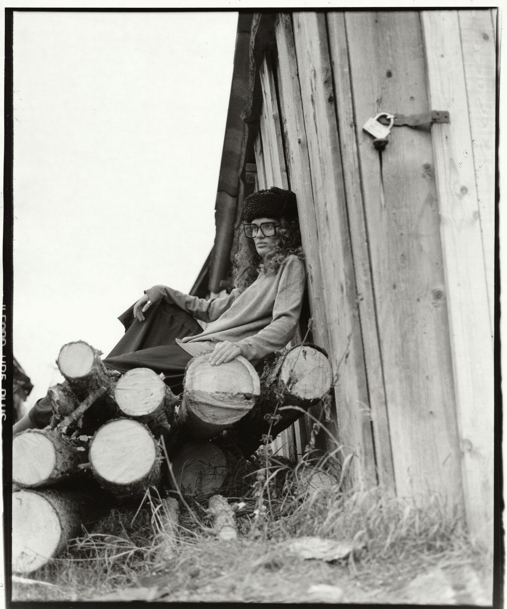 Woman in beret and glasses sits on stacked logs against wooden building with film rebate visible.