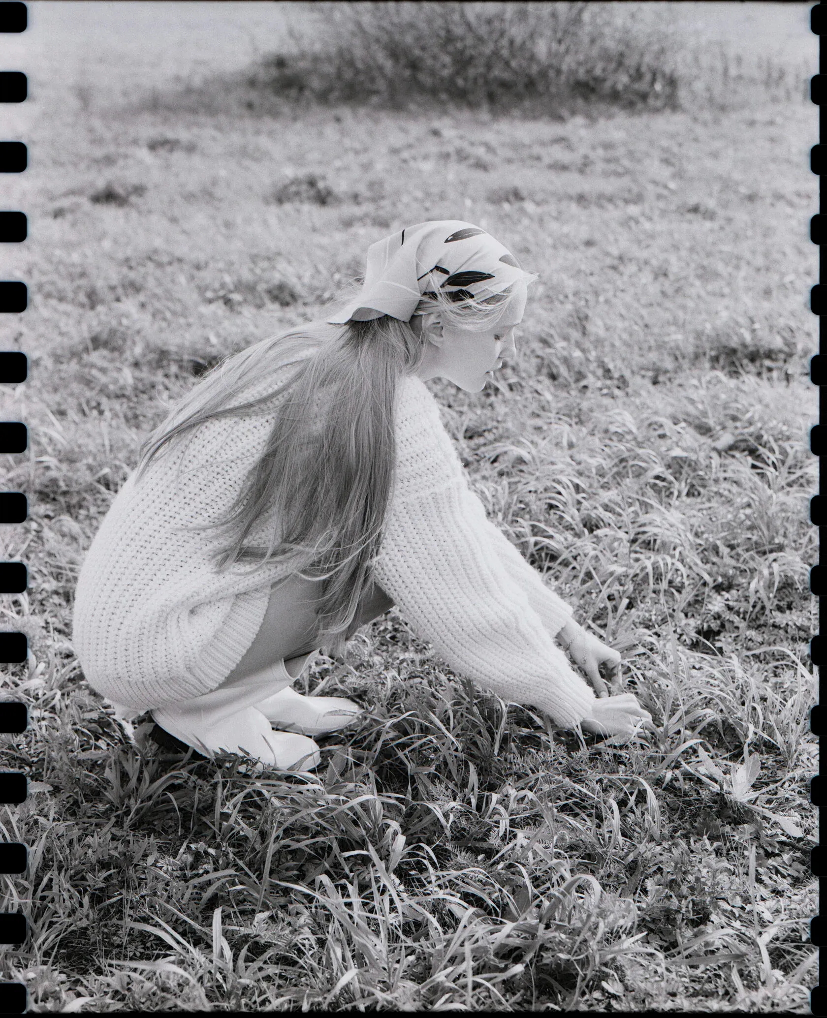 Woman in headscarf and sweater crouches in field with visible sprocket holes.