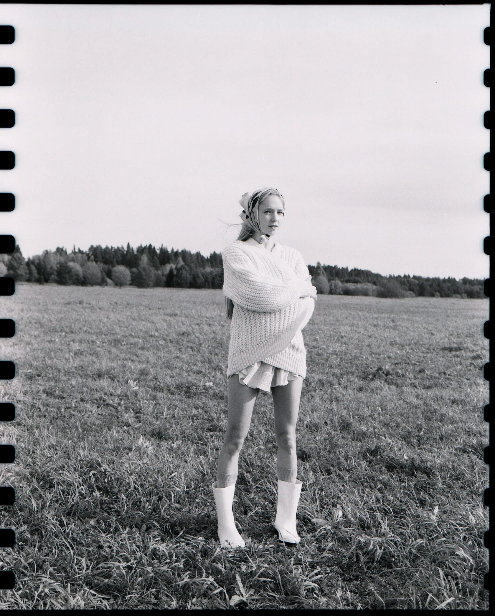 Woman in oversized white sweater stands in field with forest background and sprocket holes visible.