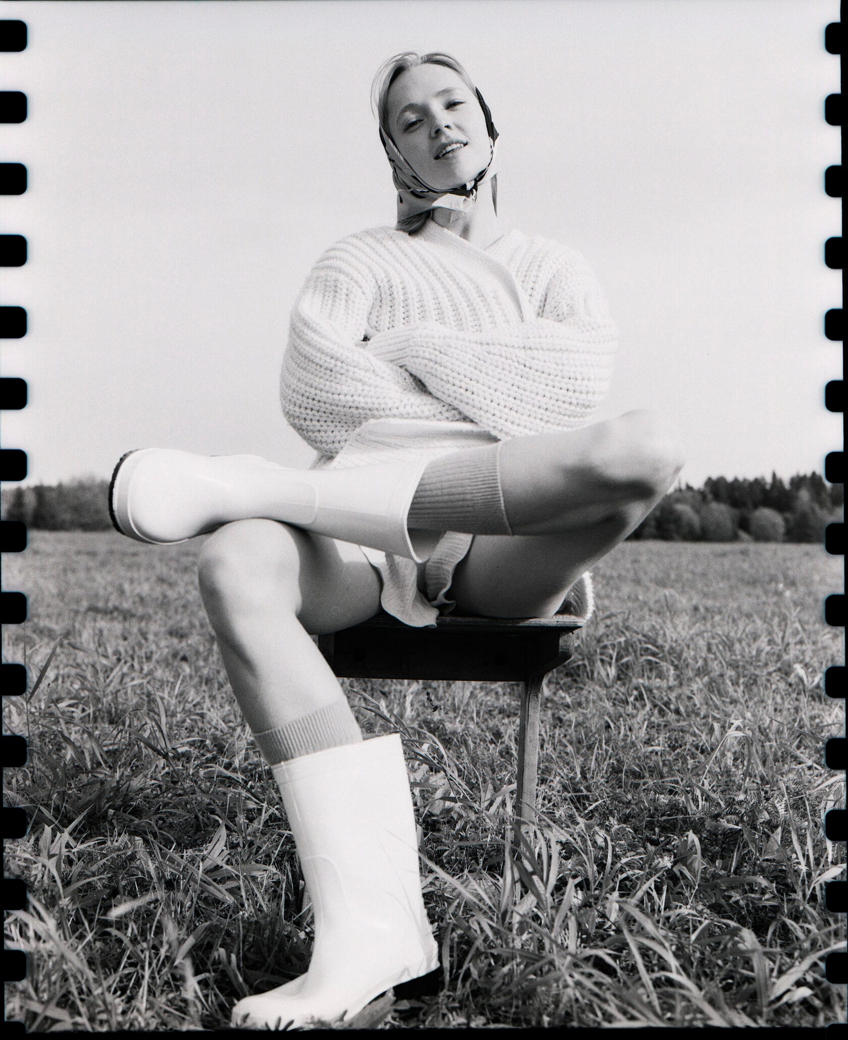 Woman in white sweater sits on stool in field with legs extended and sprocket holes visible.