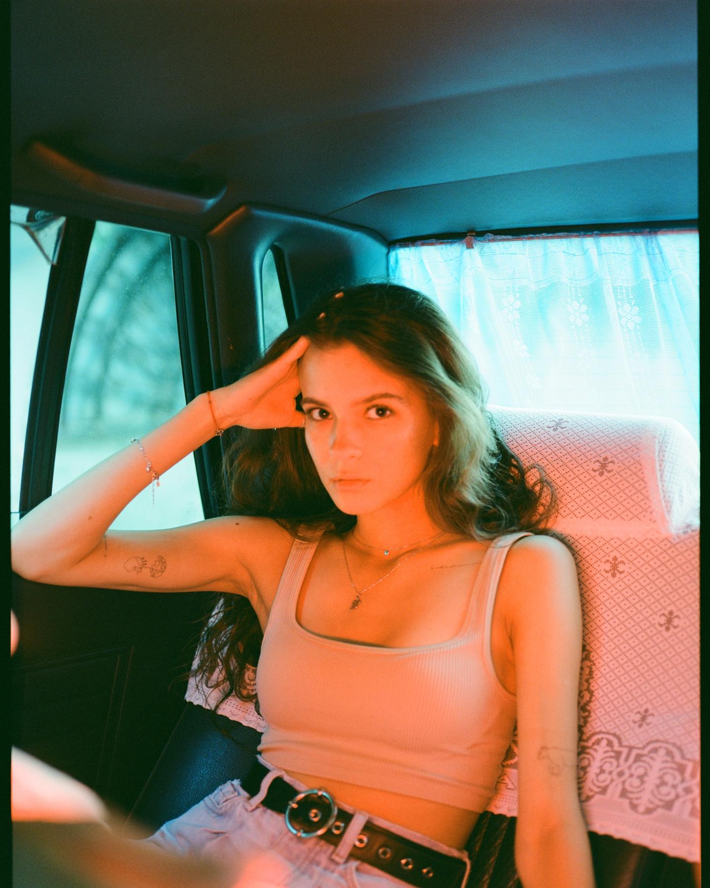 Woman in tank top saluting inside vintage van with lace curtains and warm orange light.