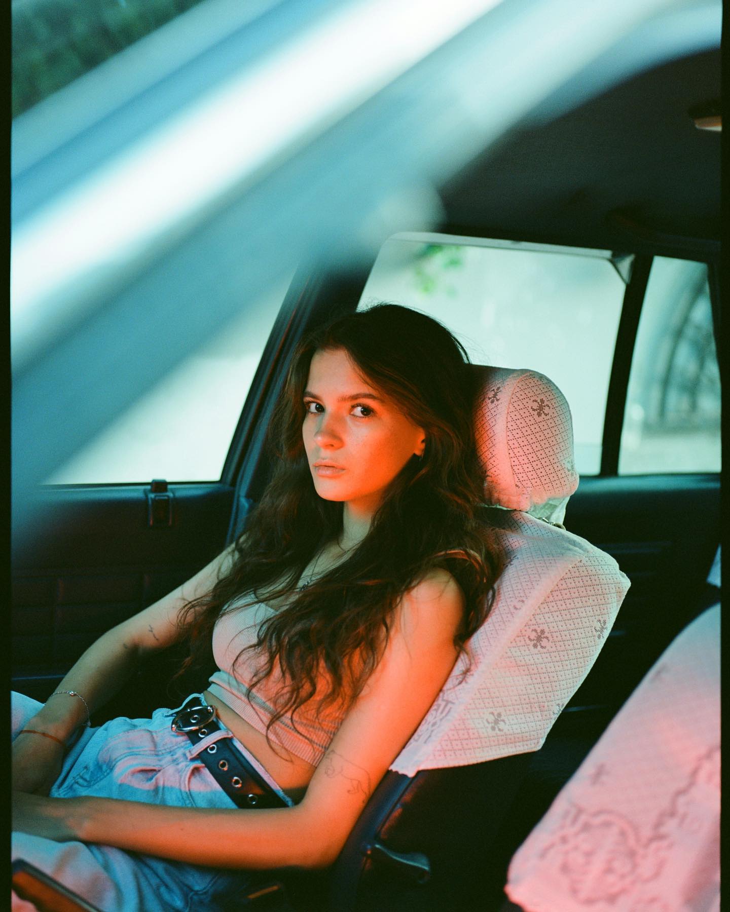 Portrait of model with long dark hair sitting in car backseat with vintage lace headrest.