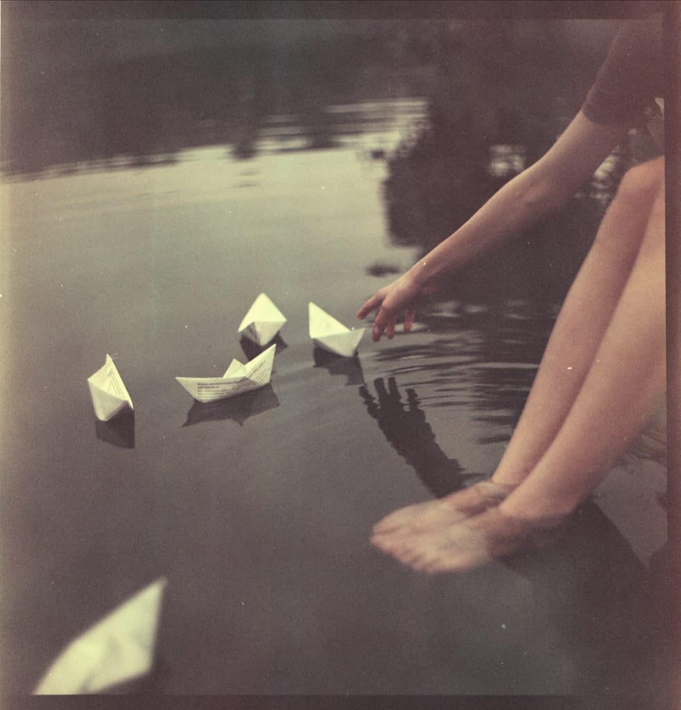 Hands placing paper boats into still water with motion blur creating dreamy atmosphere.