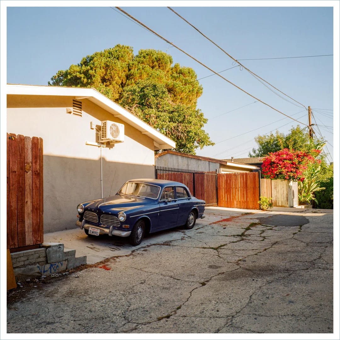 Vintage blue Volvo Amazon parked in driveway beside white stucco house with red flowering tree.