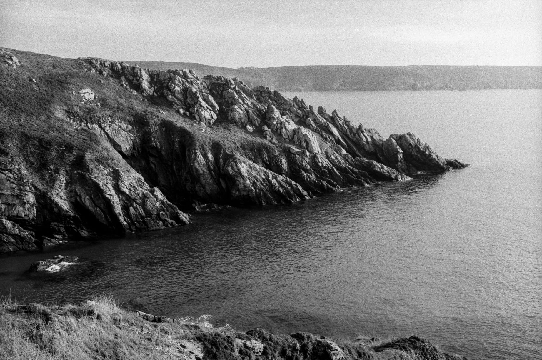 Jagged coastal rocks rise from calm water with grassy slopes above.