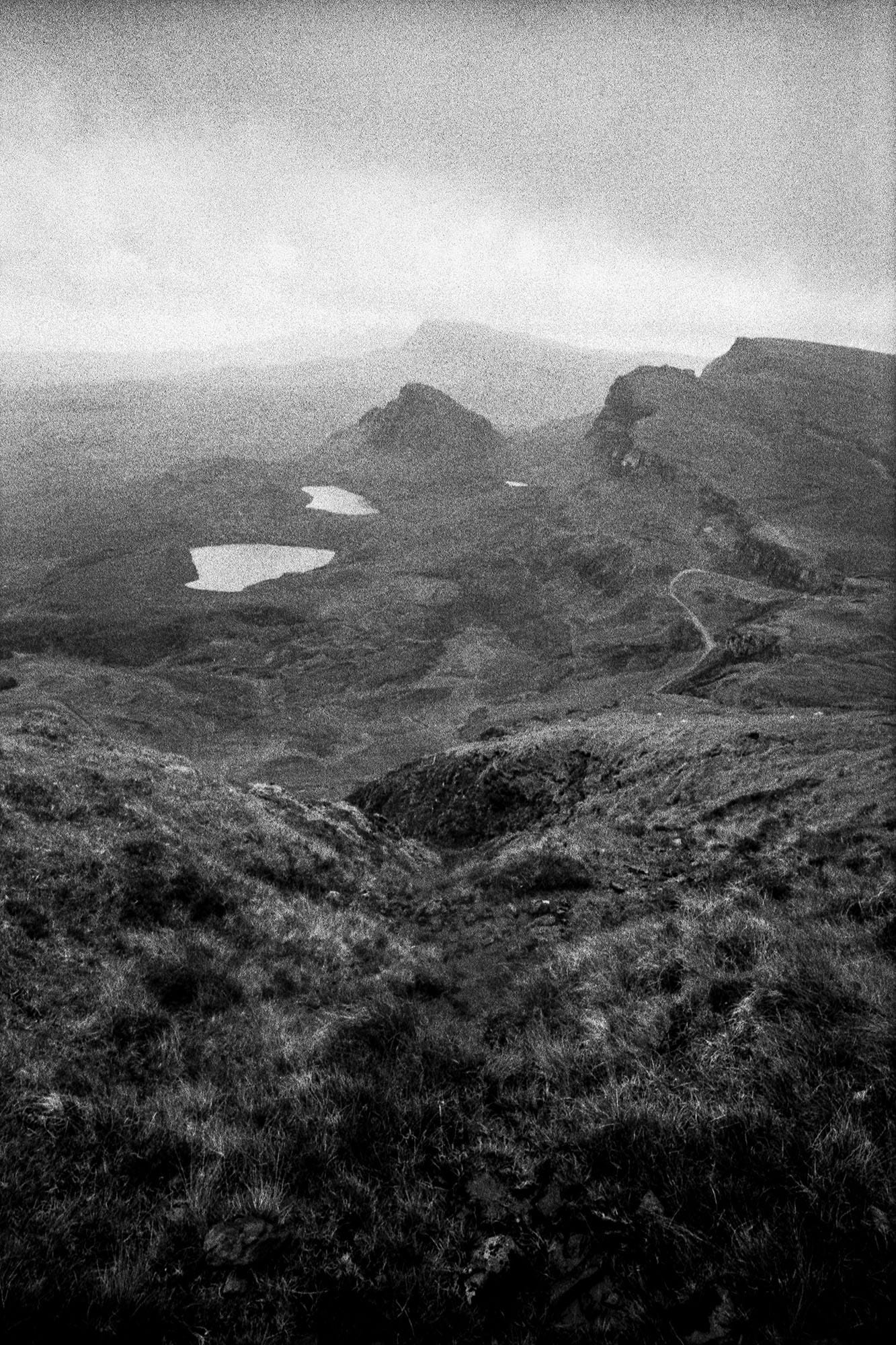 Mountain lakes visible through grainy mist with foreground highland vegetation.