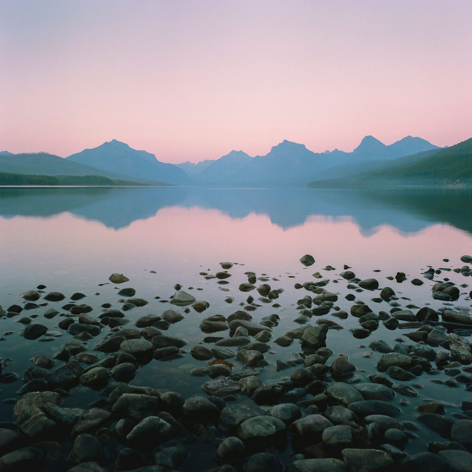 Pink dawn sky reflects in calm mountain lake with rocky foreground.