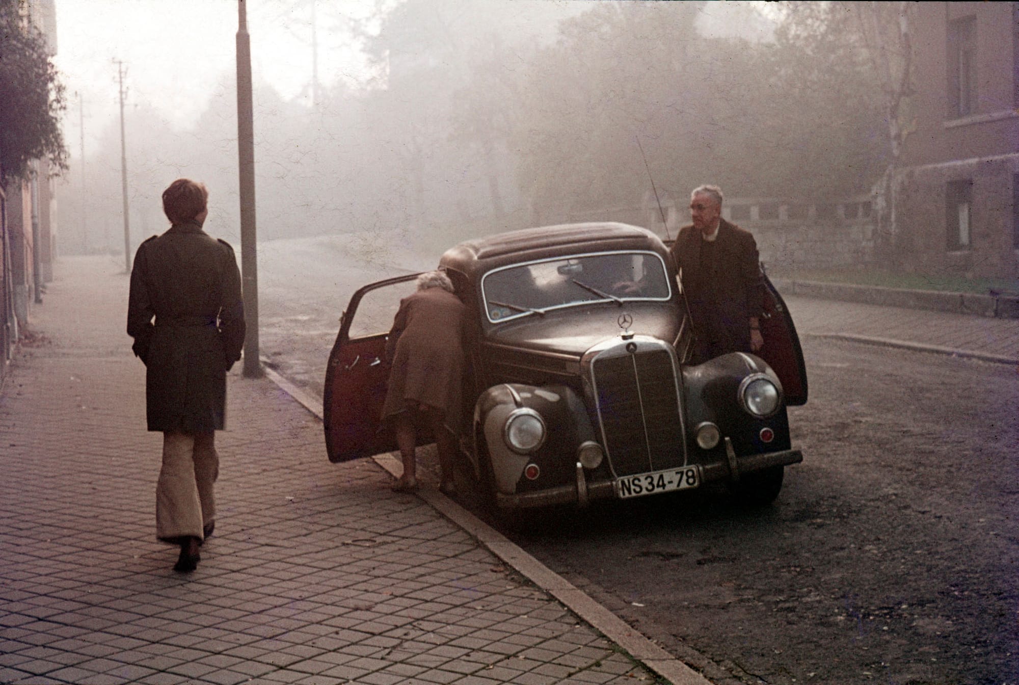 Vintage street photography of person walking past weathered wall and classic Mercedes.