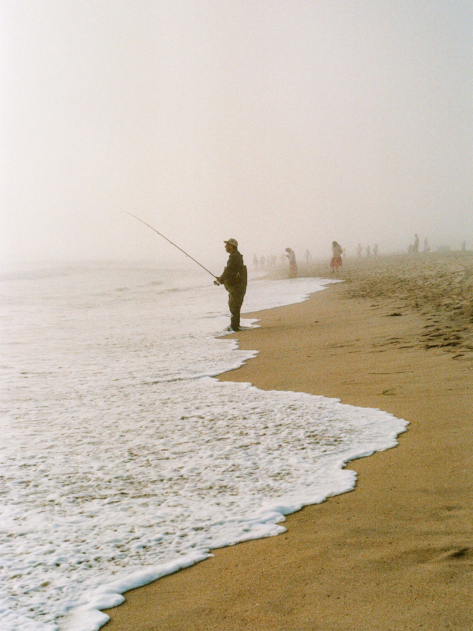 Lone fisherman stands in surf on foggy beach with distant figures visible.