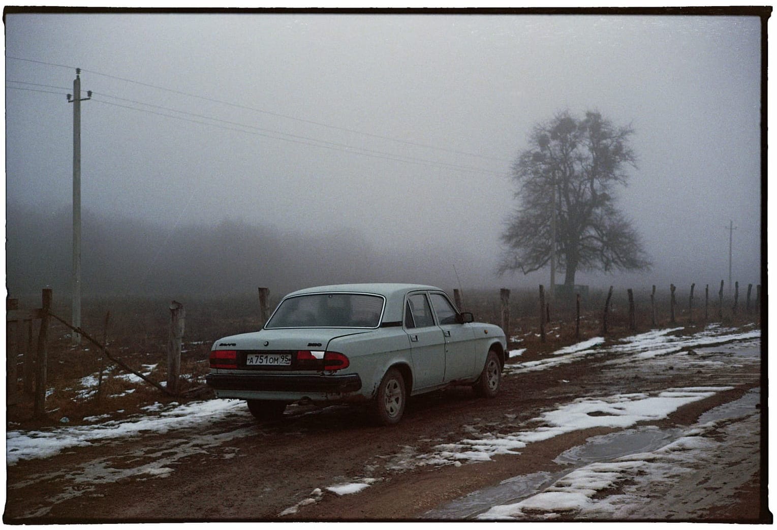 Light blue vintage sedan parked on muddy rural road with wooden fence and bare tree in dense fog.