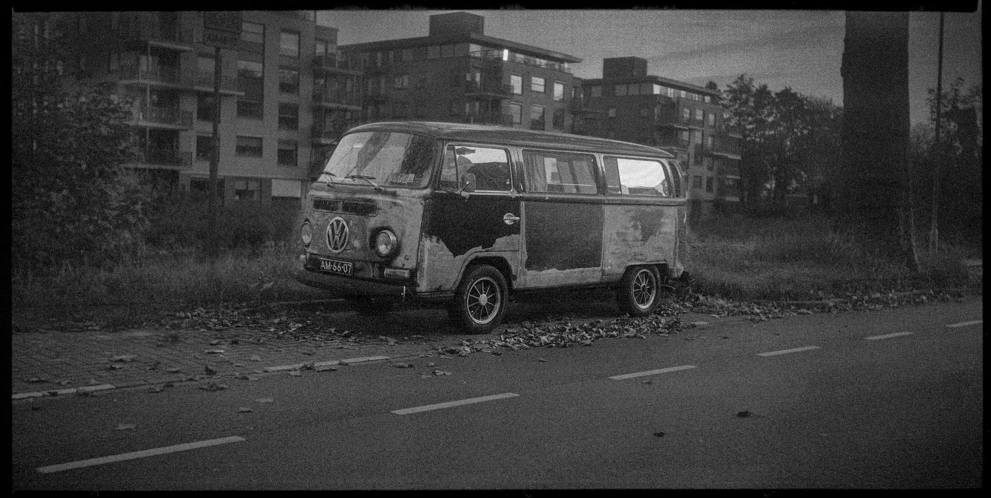 Black and white abandoned rusty Volkswagen van parked in empty lot near apartment buildings.
