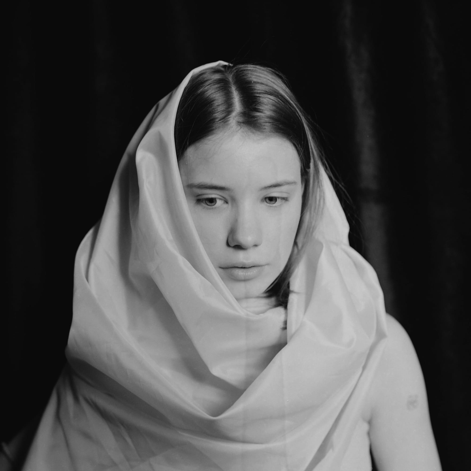 Black and white portrait of a girl wrapped in translucent white fabric against dark background.