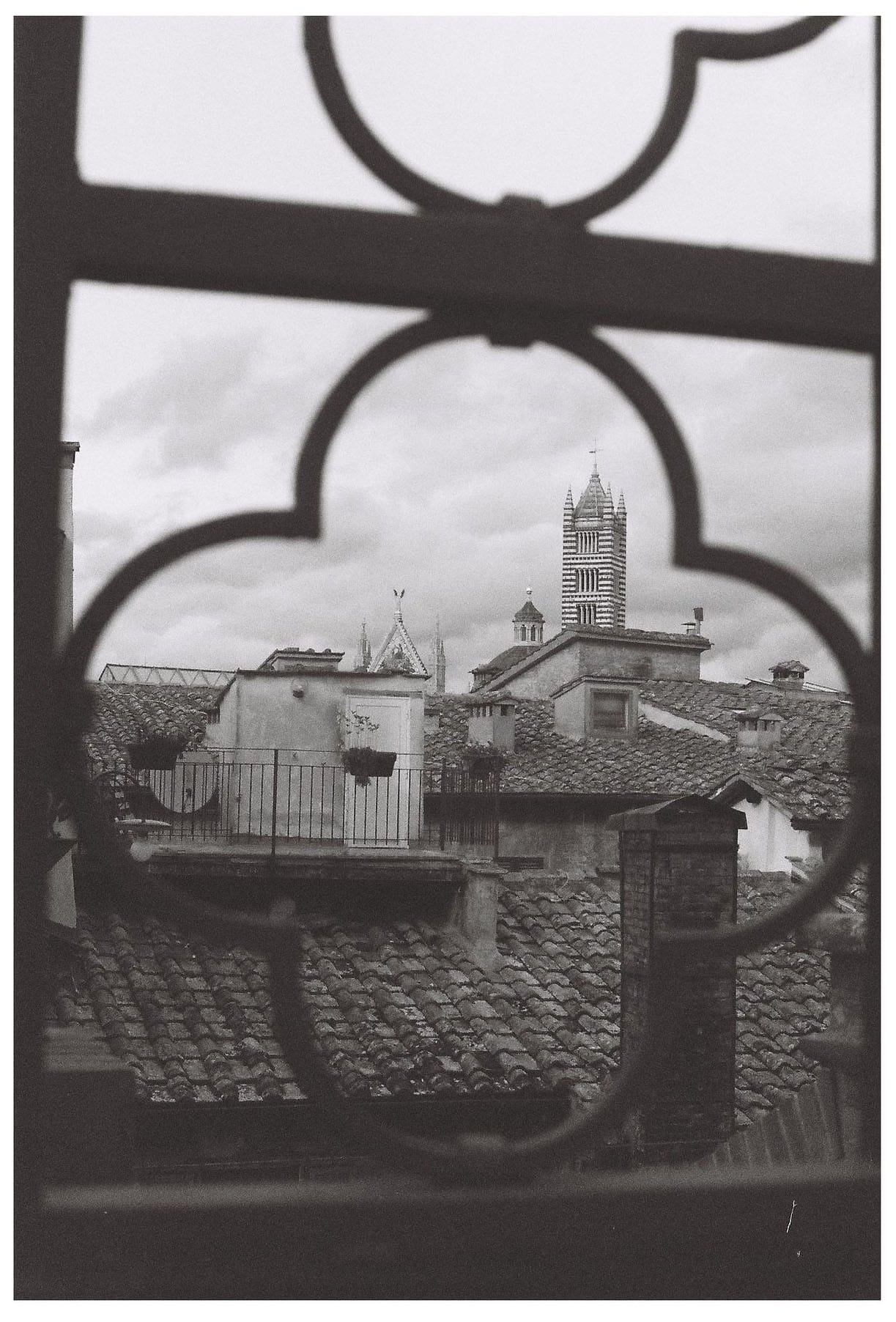 Black and white view of cathedral bell tower through ornate window frame with rooftops.