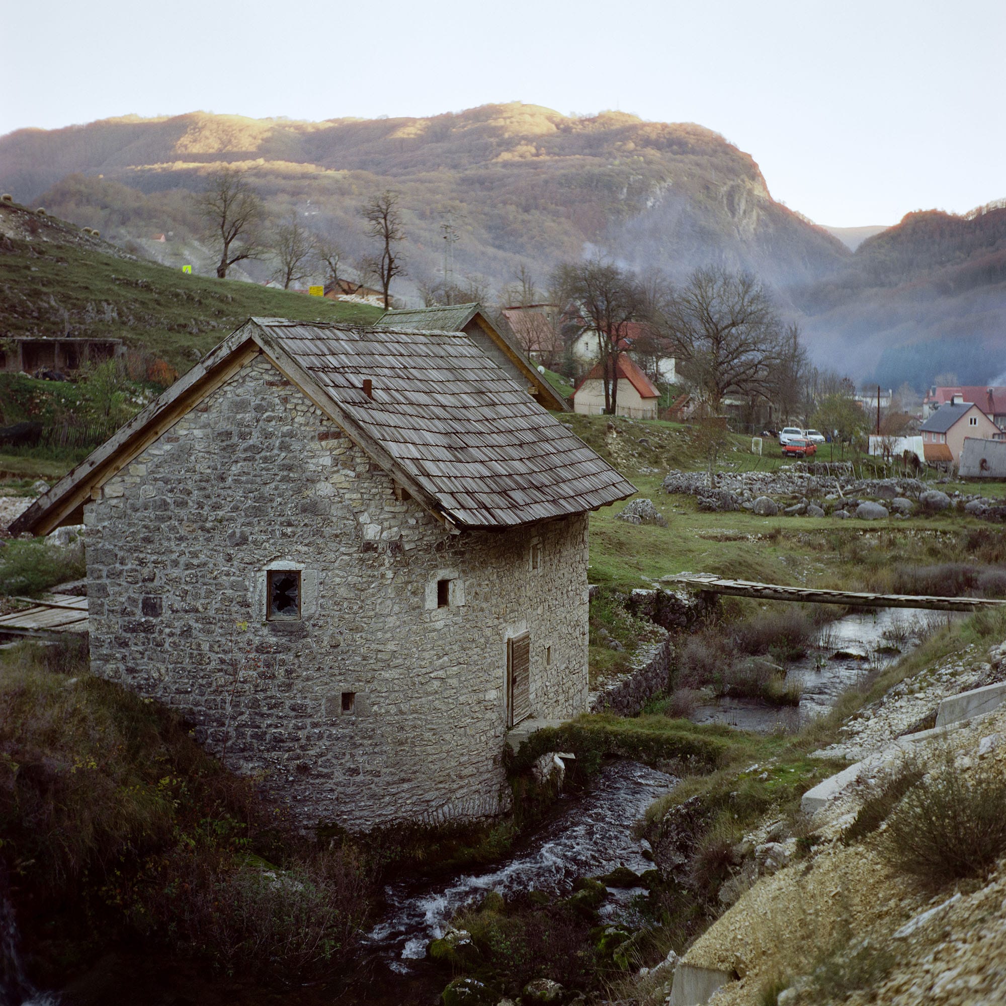 Old stone watermill with wooden roof beside stream in mountain village at dusk.
