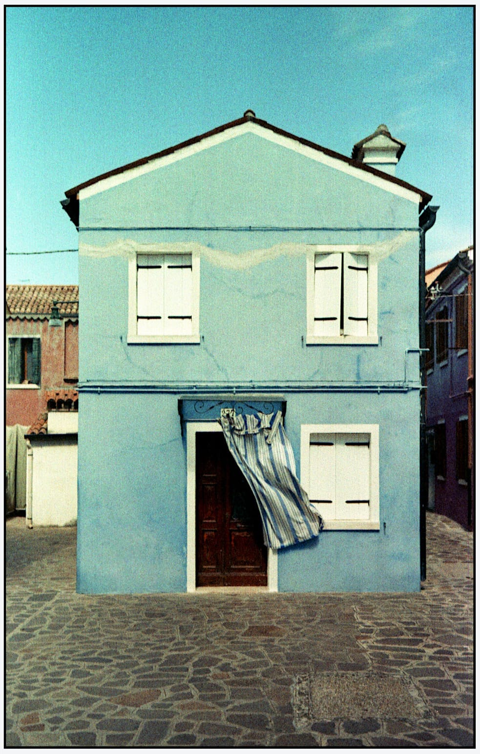Turquoise house facade with white shutters and striped curtain in doorway.