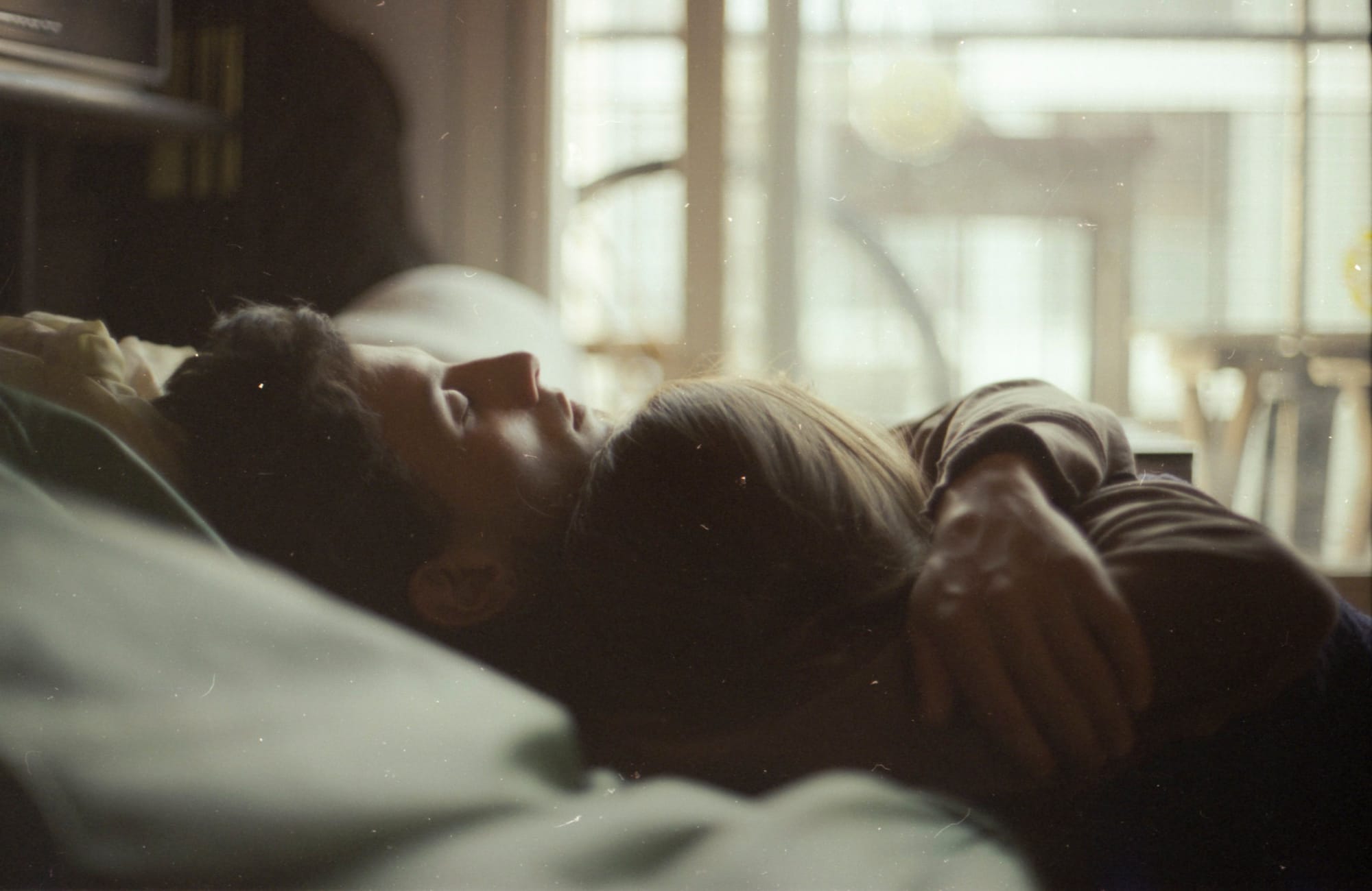 Intimate couple portrait lying in bed near window with soft natural light and film grain.