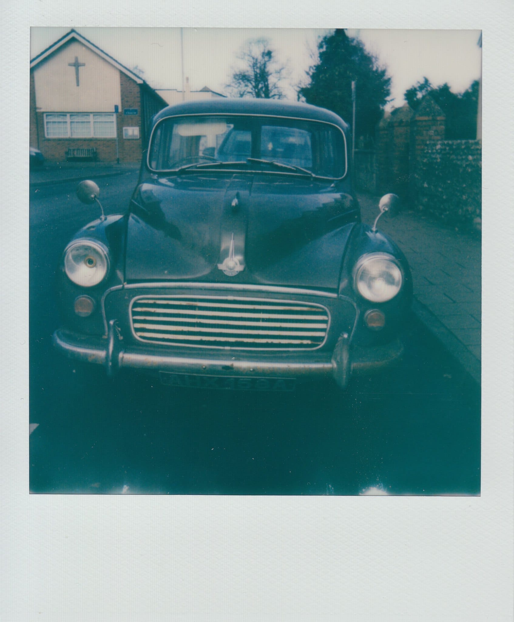 Weathered Morris Minor front view with church building behind in blue-toned Polaroid.