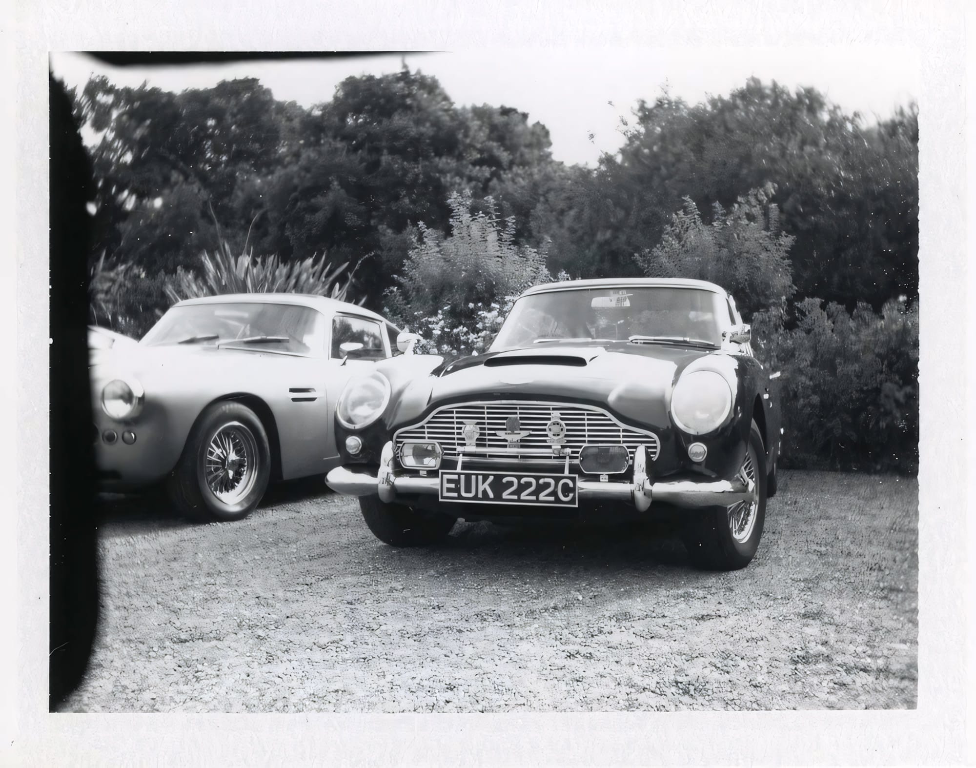Two Aston Martin DB models at car show with trees in background black and white instant film.