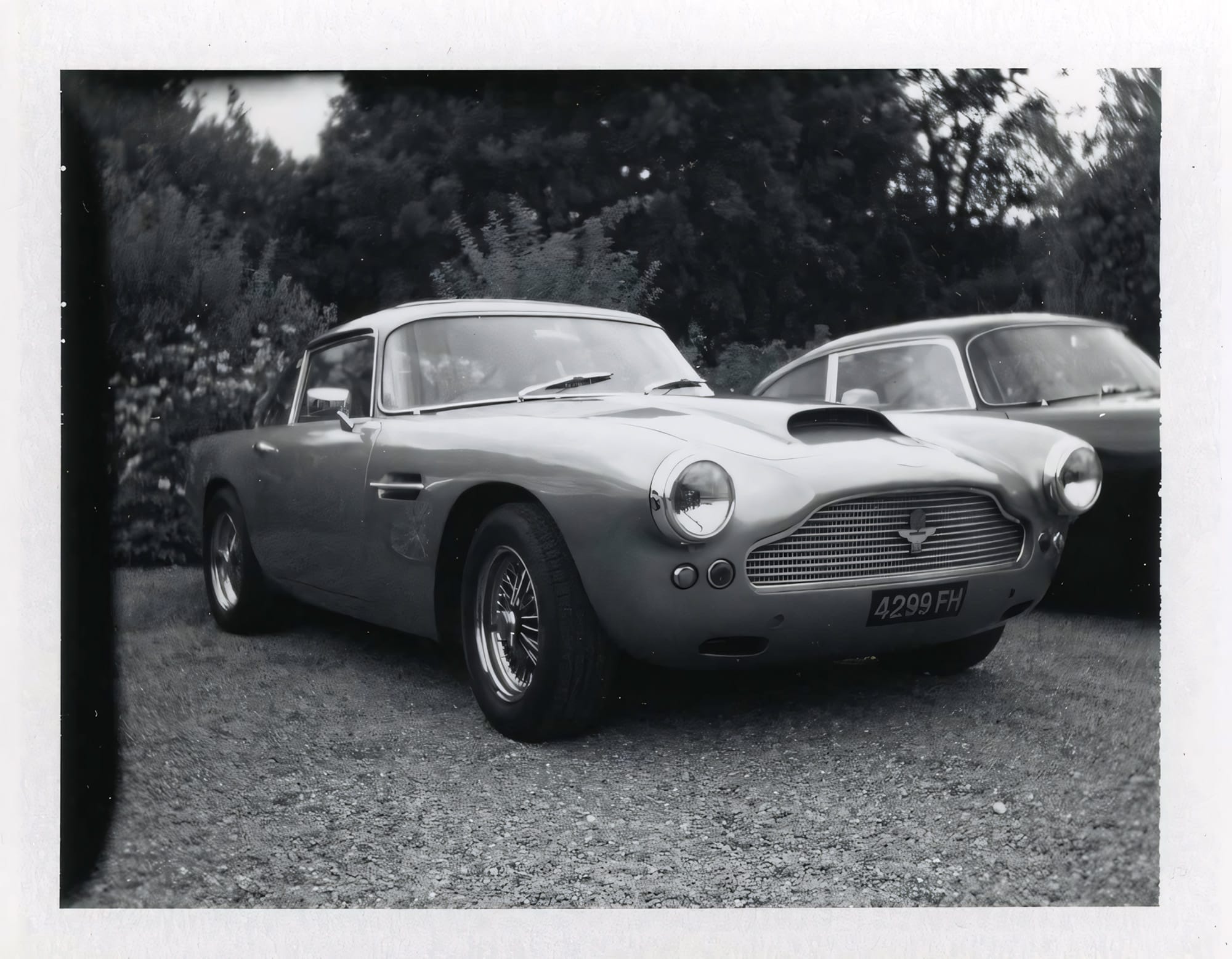 Silver Aston Martin DB4 at outdoor car show on gravel with trees behind black and white Polaroid.