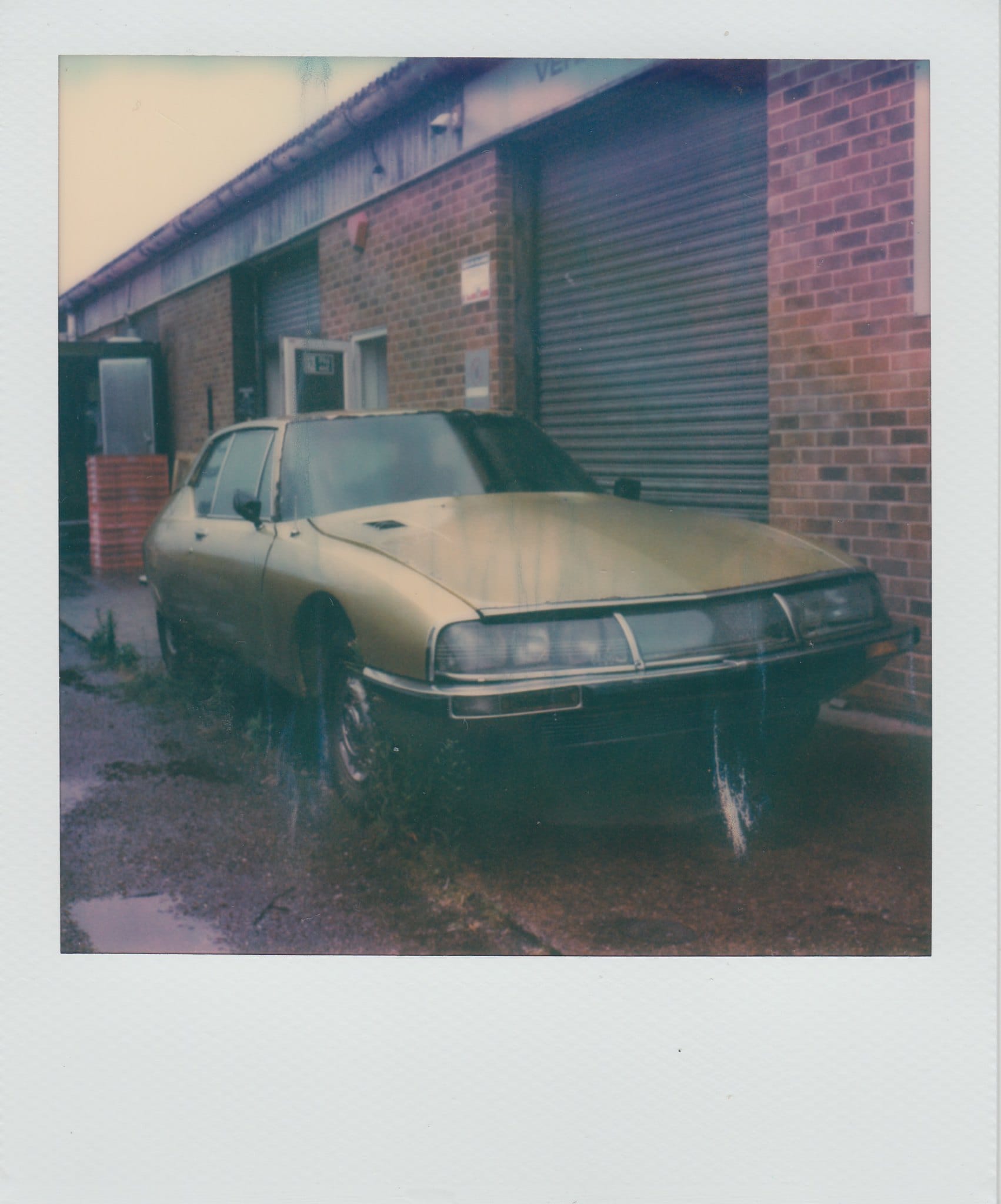 Weathered Citroën SM beside brick warehouse with corrugated door in greenish Polaroid.