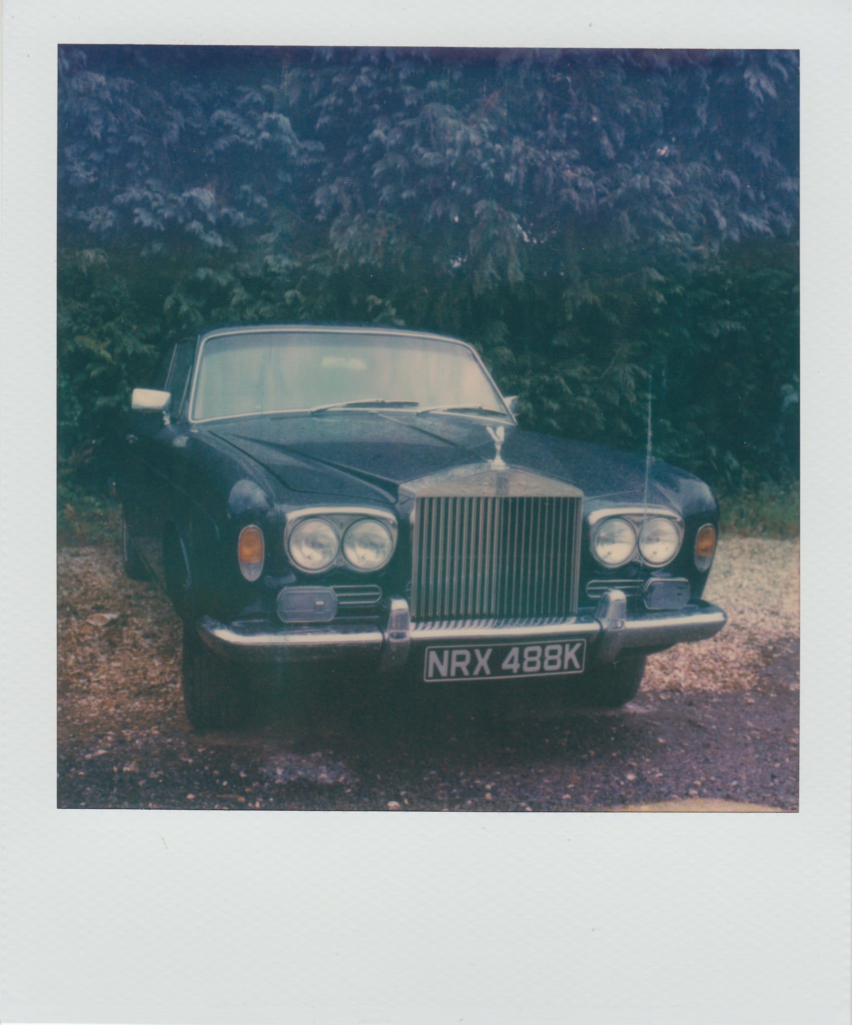 Dark Rolls-Royce on gravel driveway with trees in background faded Polaroid.
