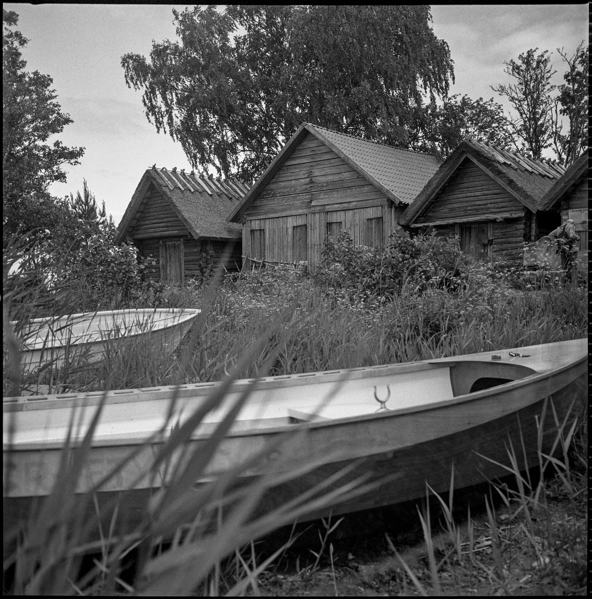 White boats rest in overgrown grass below weathered wooden buildings.