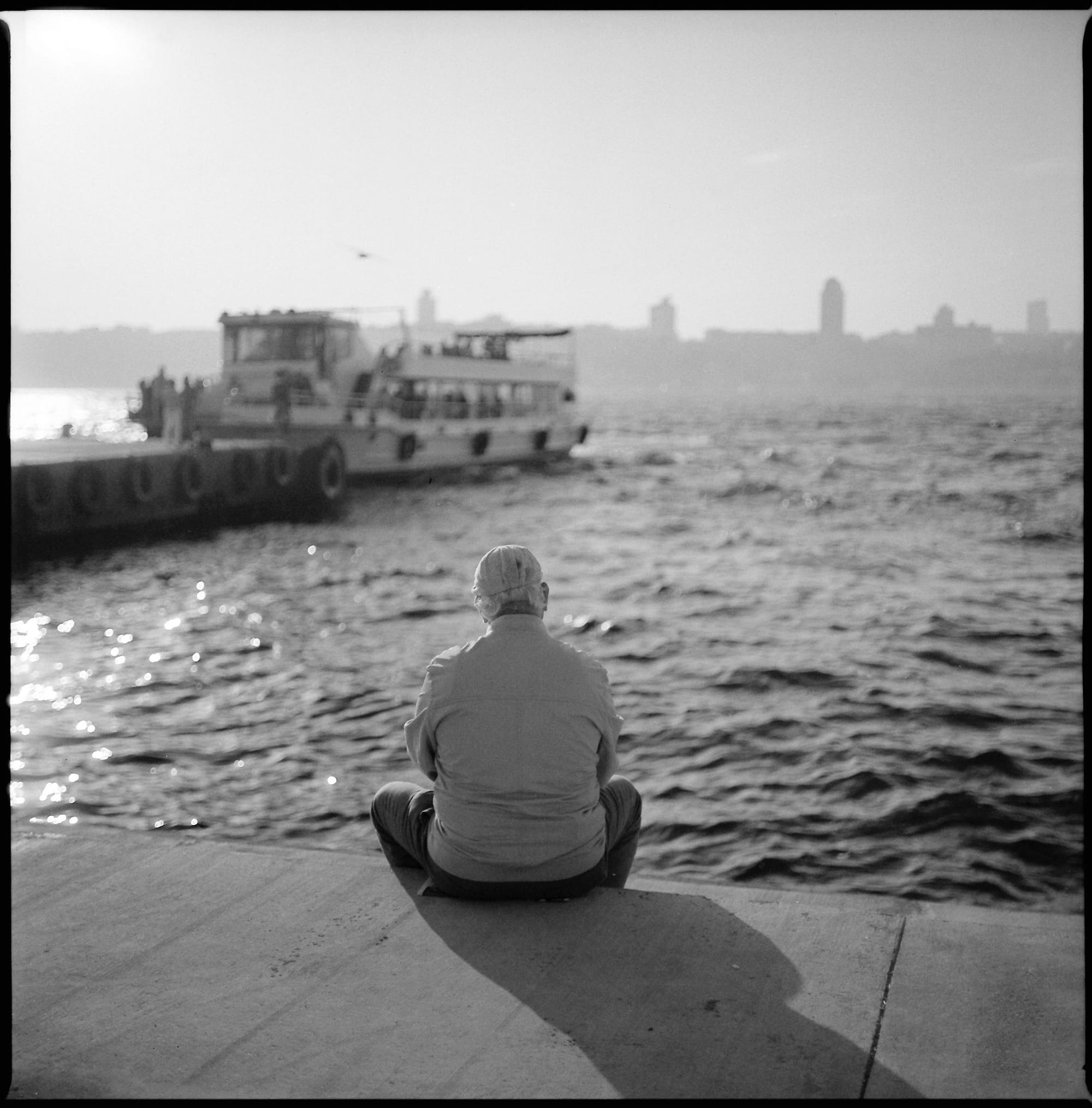 Man sits alone on waterfront looking toward ferry and distant buildings in Istanbul.