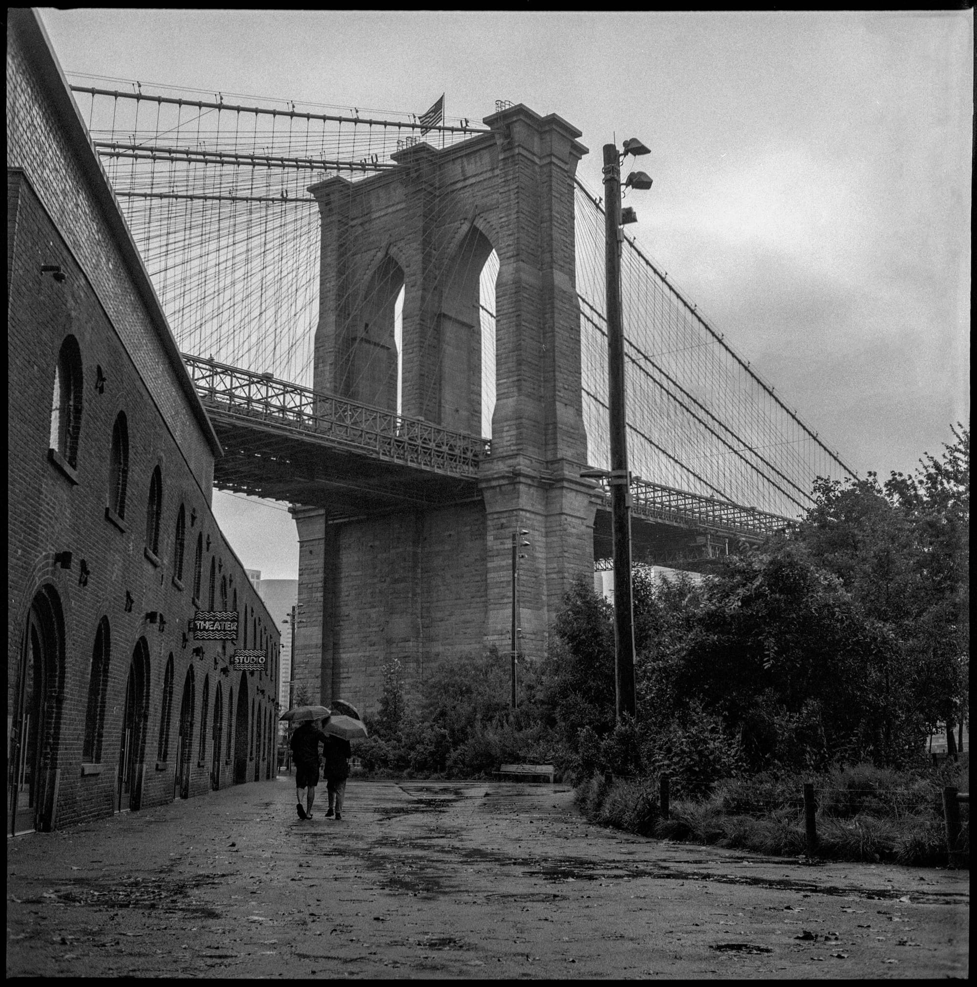 Brooklyn Bridge tower looms over wet street with two figures walking.