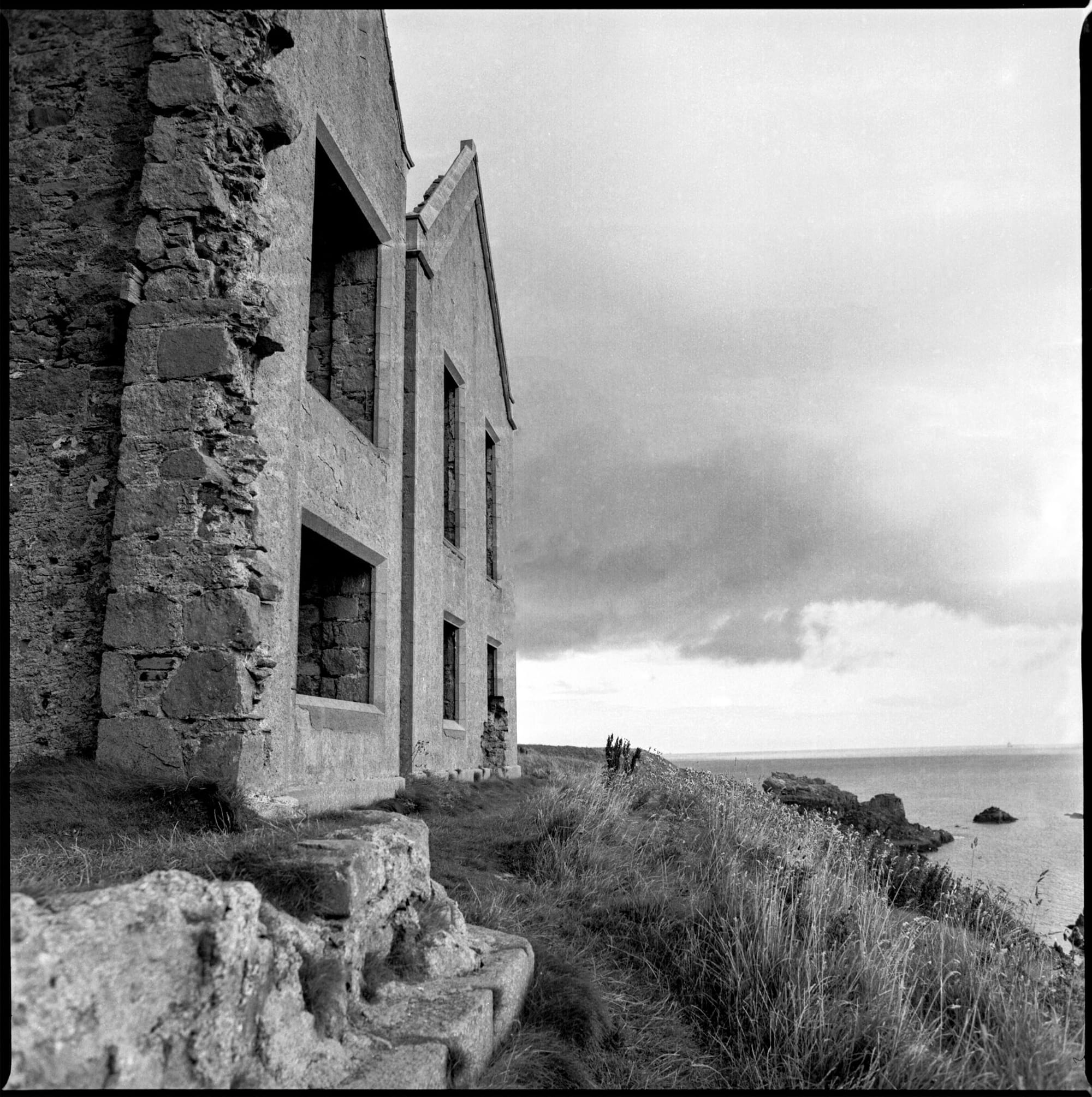 Ruined coastal building overlooks ocean from rocky cliff.
