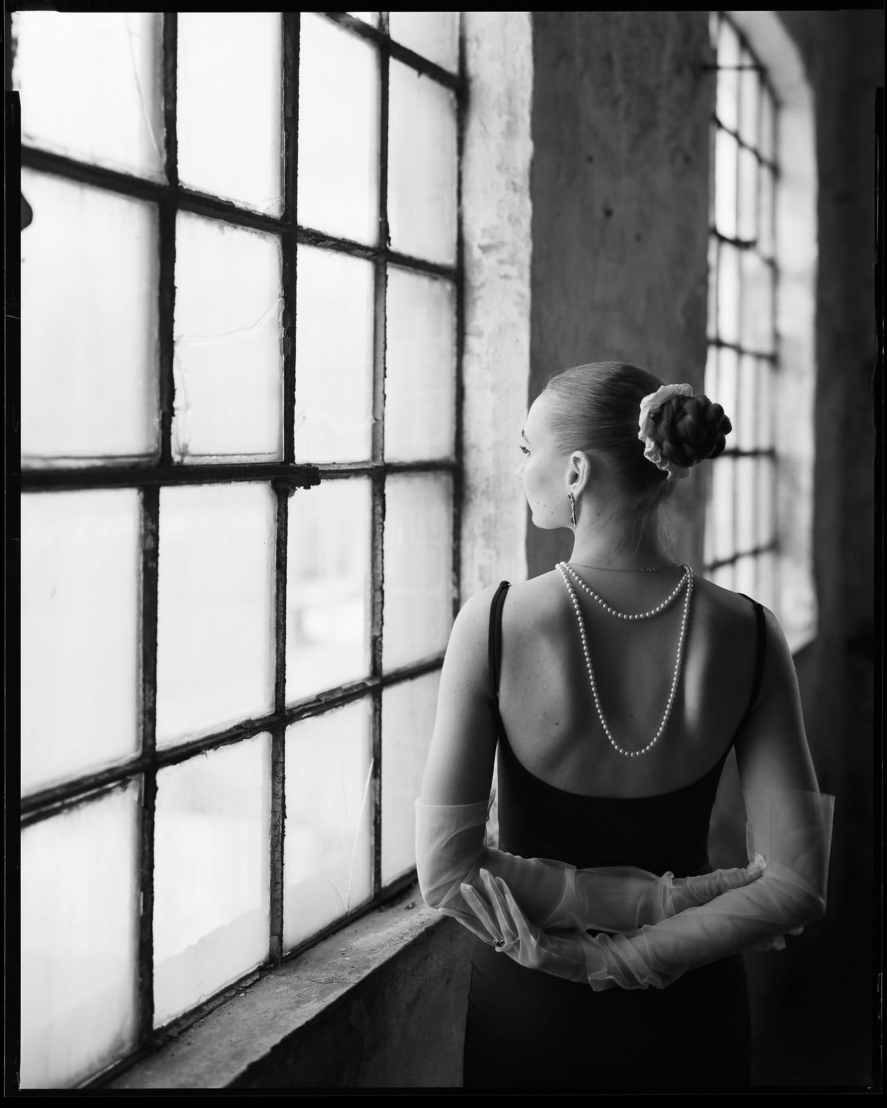 Black and white portrait of woman in dress with hair bun looking out industrial factory window.