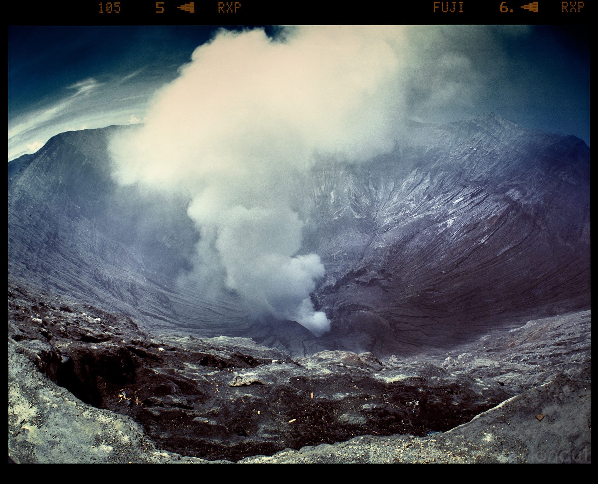 Volcanic crater releases steam plume against mountain backdrop with visible film border.
