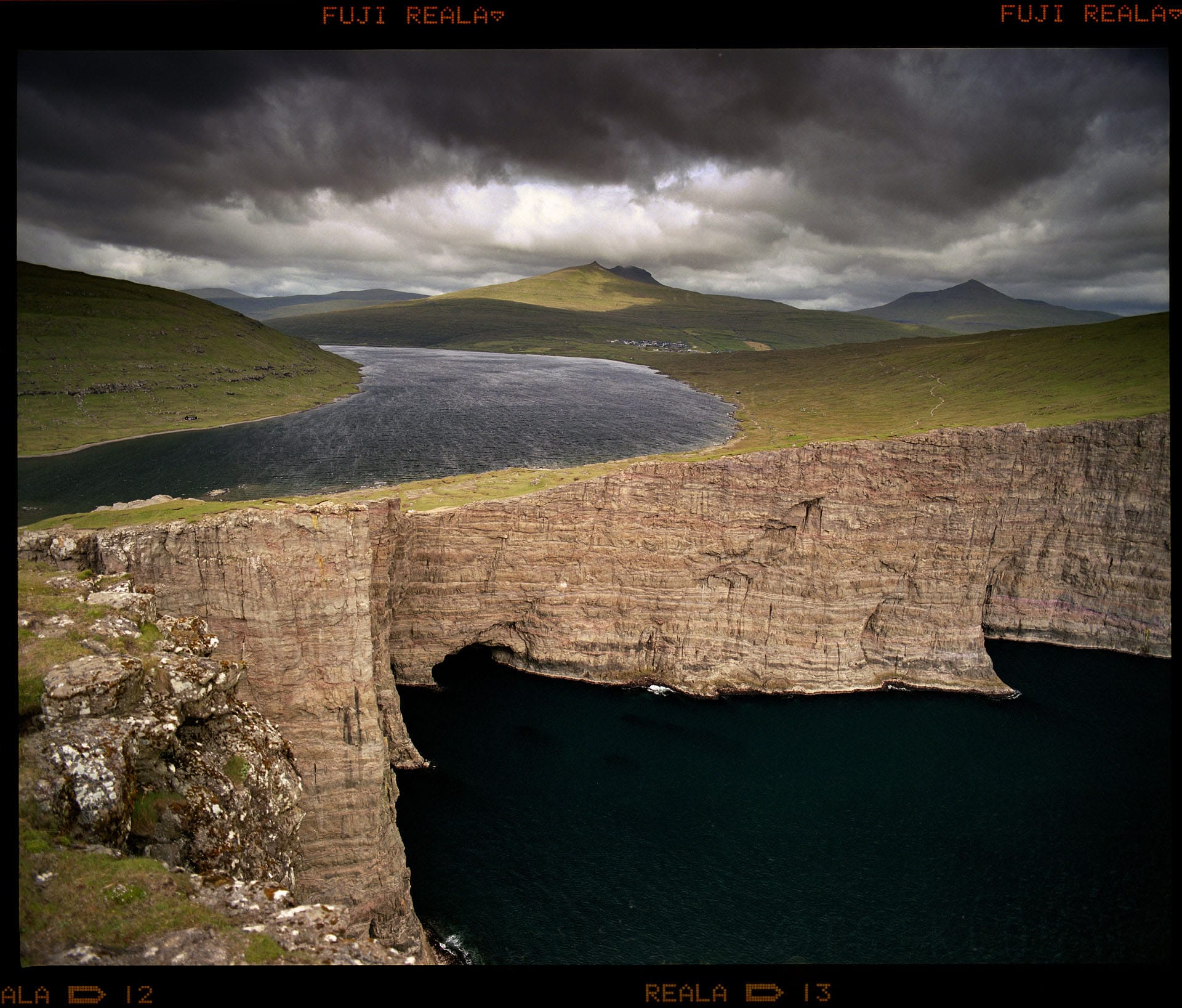 Dramatic cliffs frame turquoise lake under stormy Faroe Islands sky.