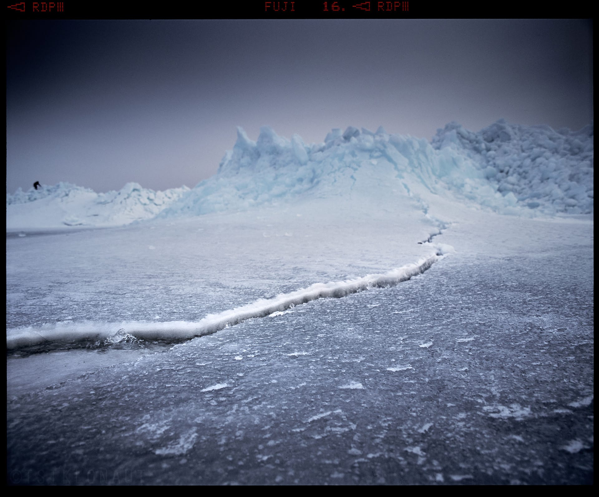 Fractured ice formations rise along frozen shoreline in blue tones.