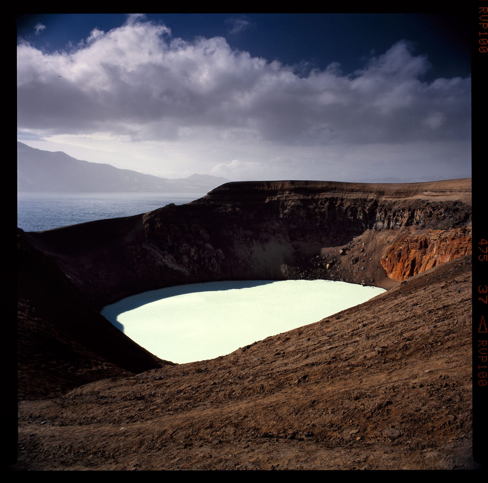 Volcanic crater lake glows turquoise against brown rock walls under storm clouds.