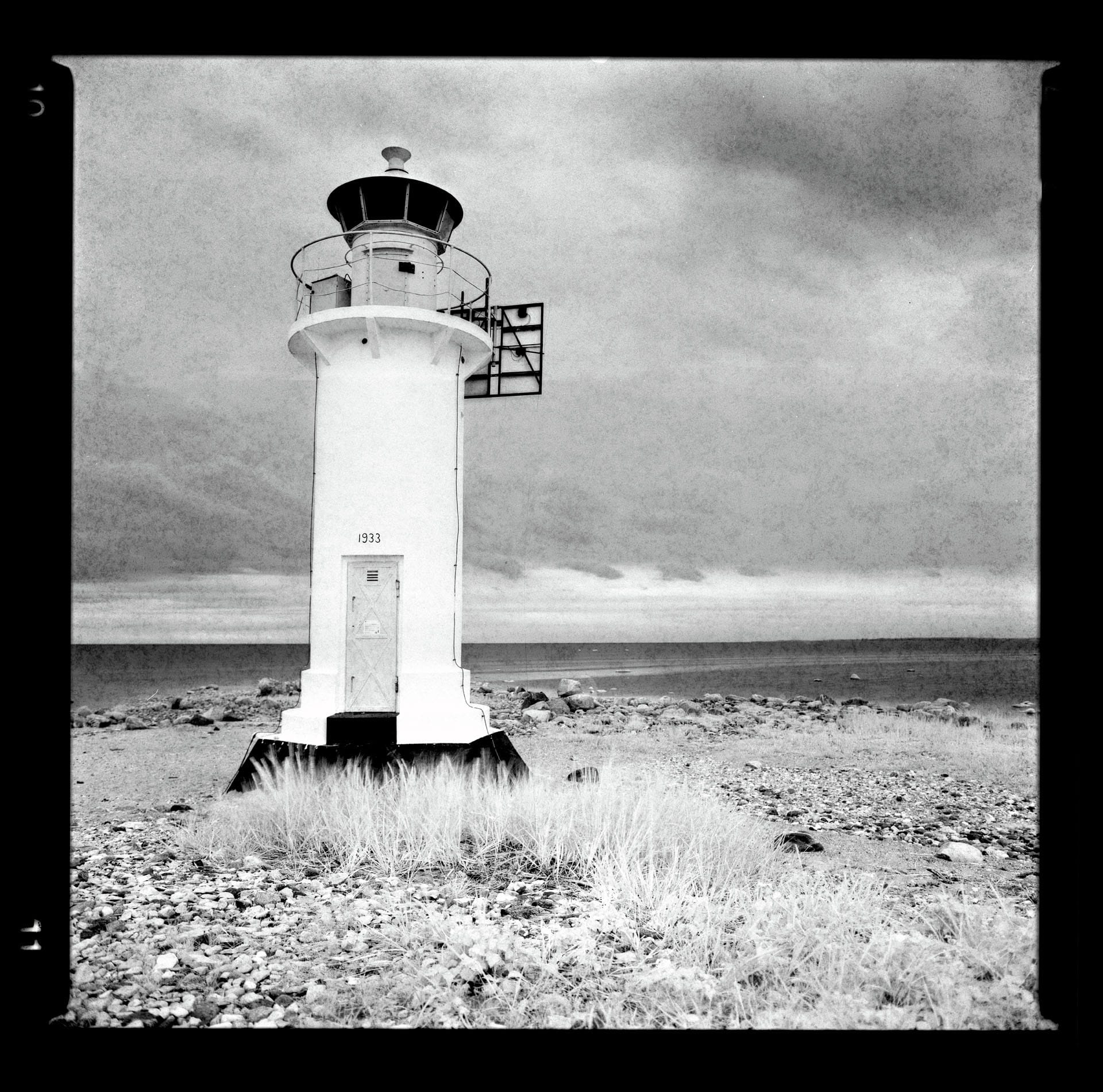 White lighthouse stands on rocky beach under threatening gray sky.