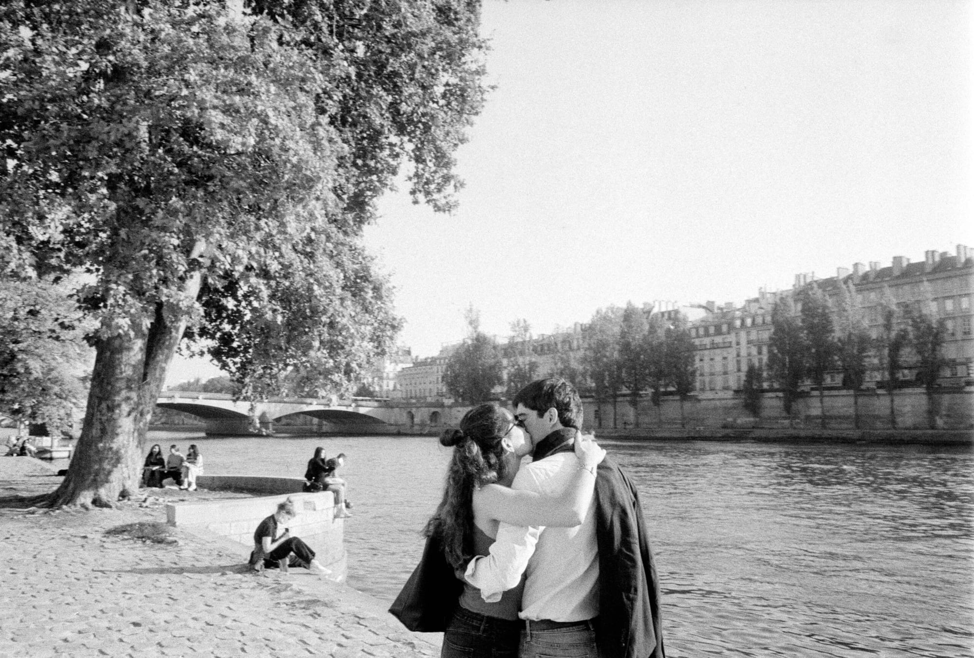 Couple embracing by Seine river with Parisian buildings and bridge visible in soft focus background.