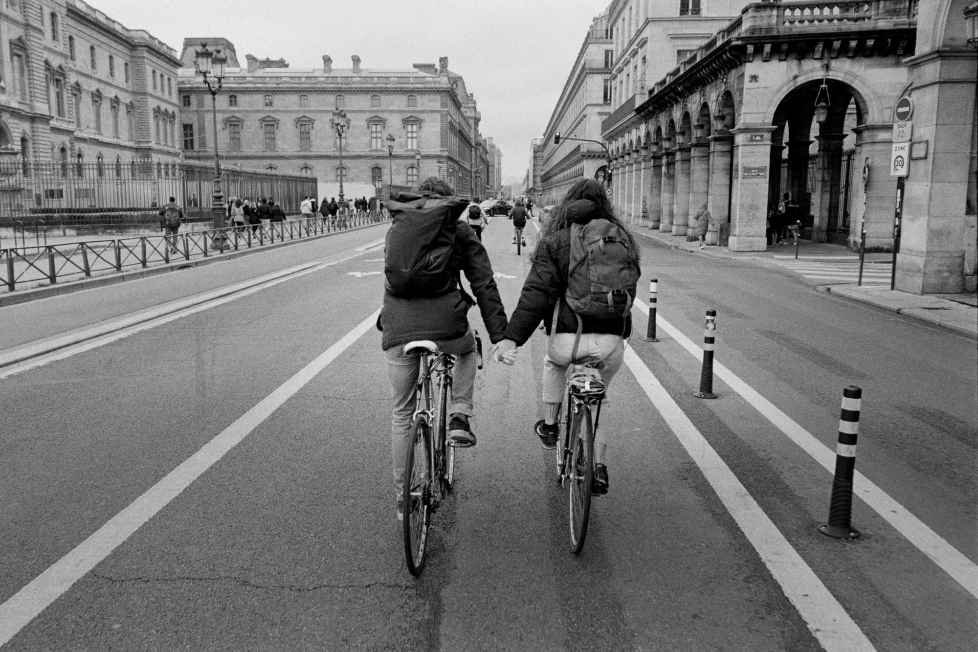 Two cyclists holding hands while riding down empty Paris street flanked by classical architecture.