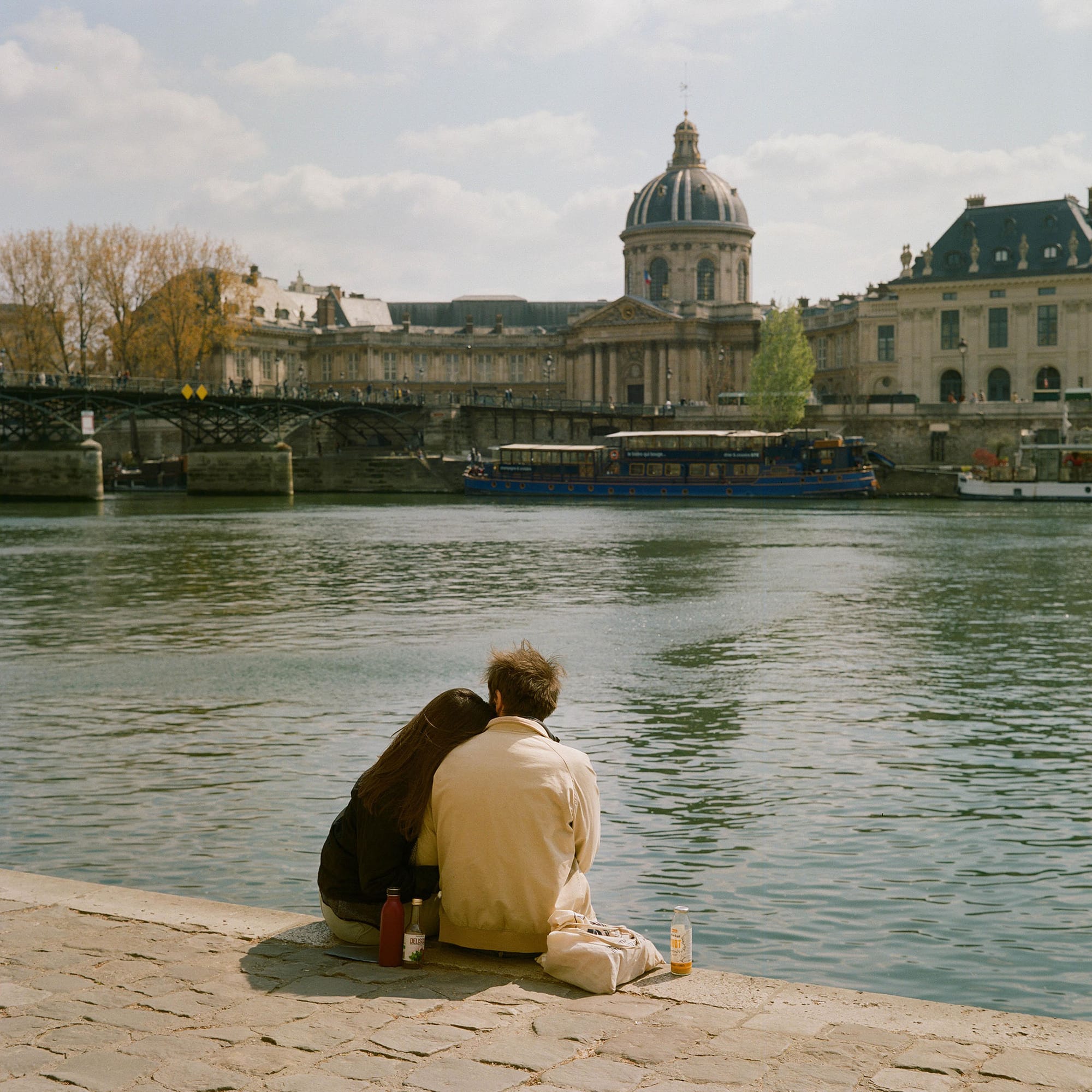 Romantic couple sitting by Seine watching boat pass with beautiful architecture in the background.