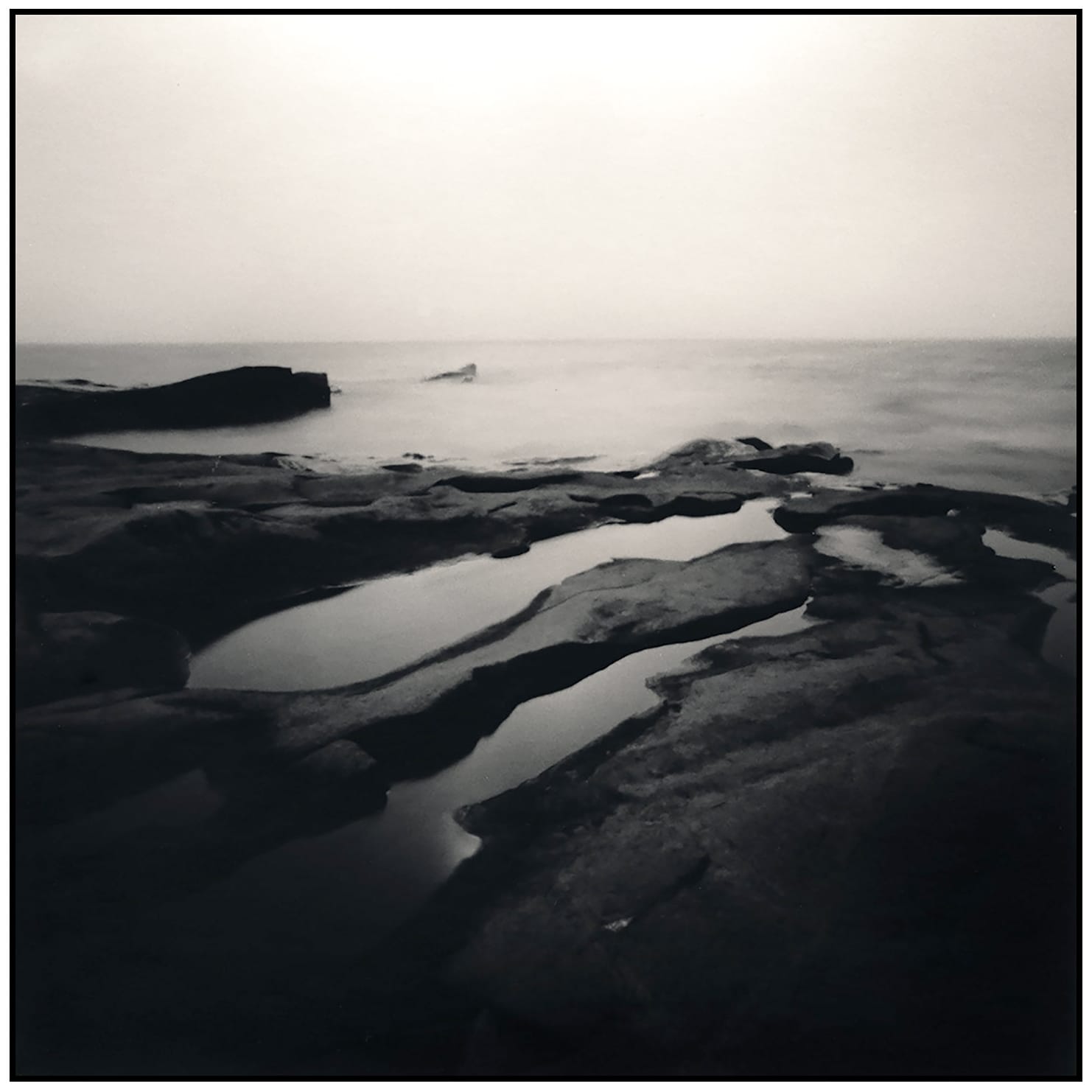 Black and white tidal pool formations creating organic shapes with smooth water reflecting overcast sky.