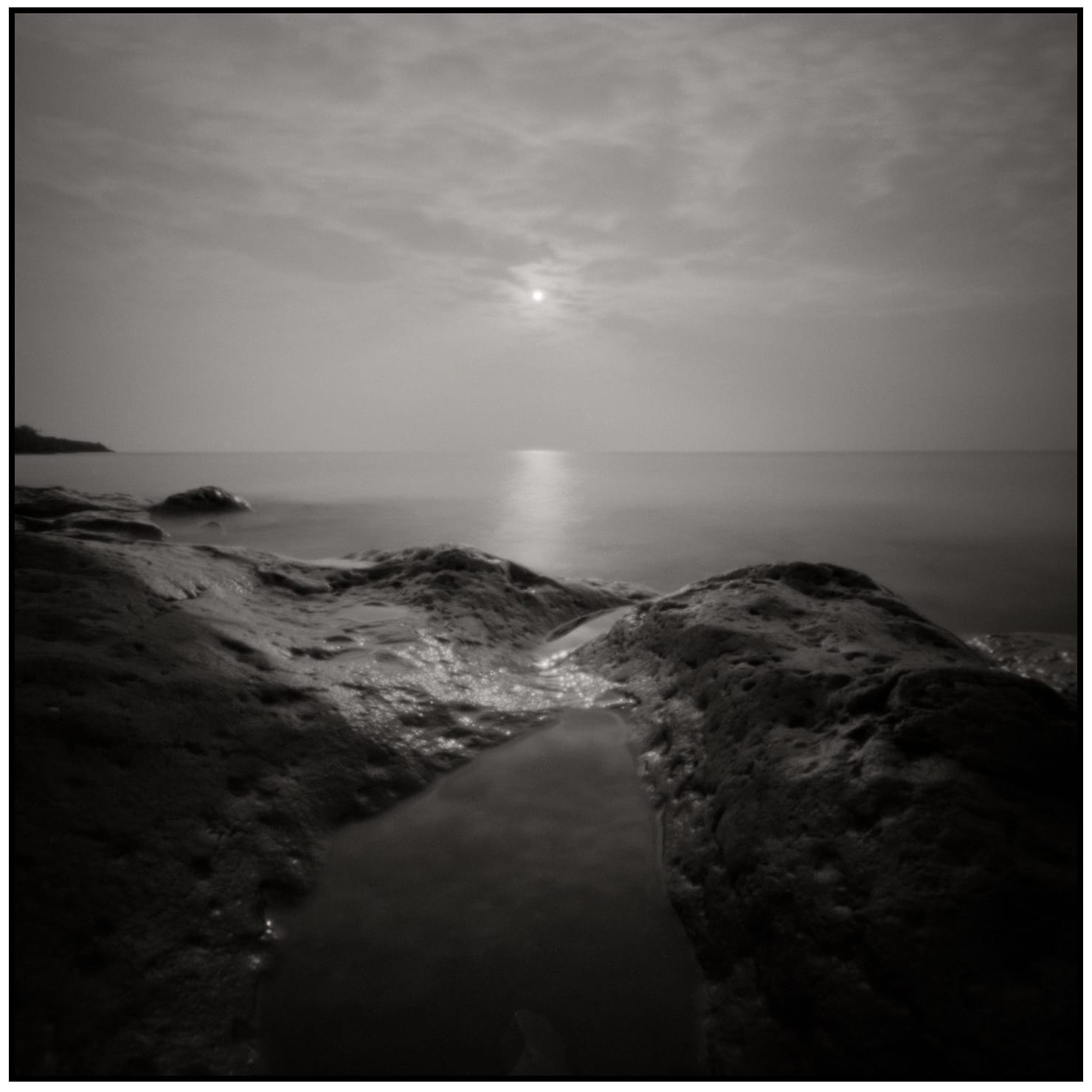 Rocky coastline at sunset with smooth water and sun creating reflection on calm sea surface.