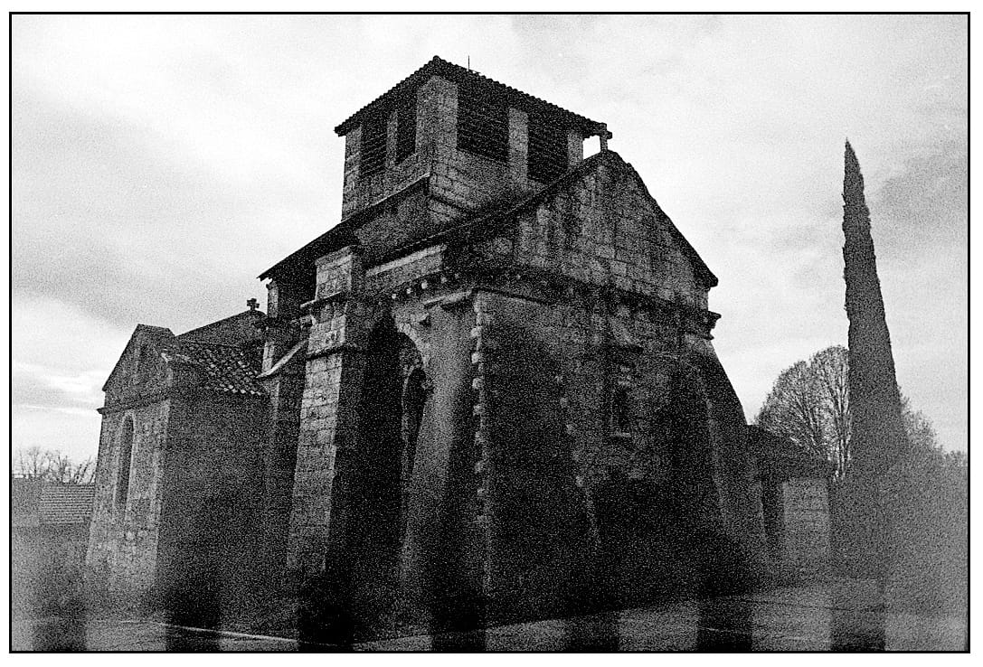 Black and white weathered stone church with bell tower and cypress tree in dramatic light.