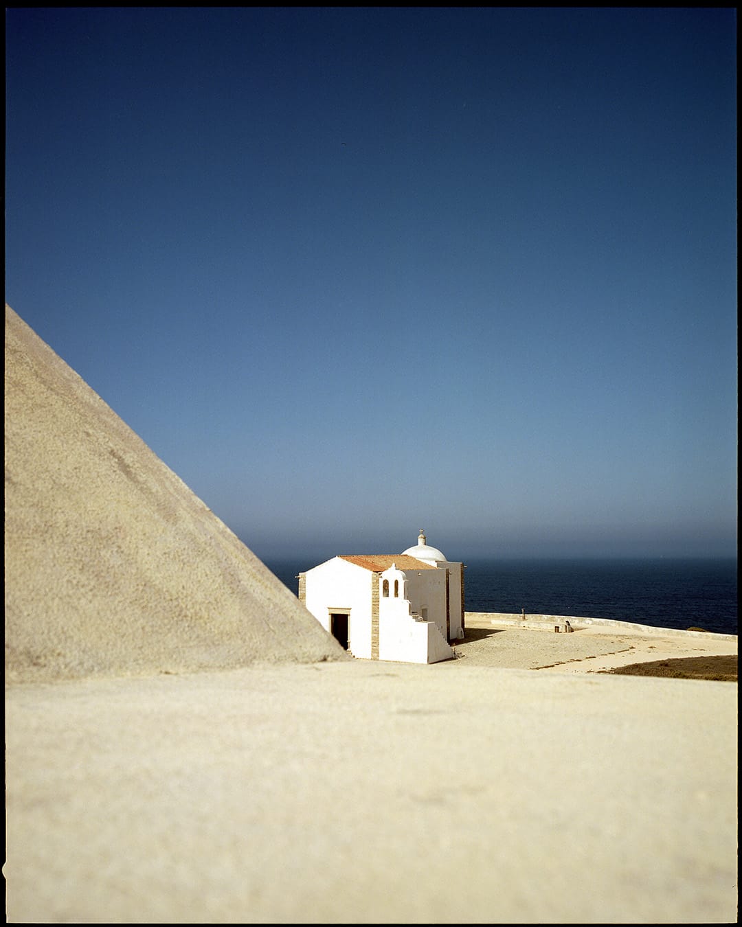 Small white Mediterranean chapel on sandy coastal cliff overlooking deep blue ocean under clear sky.