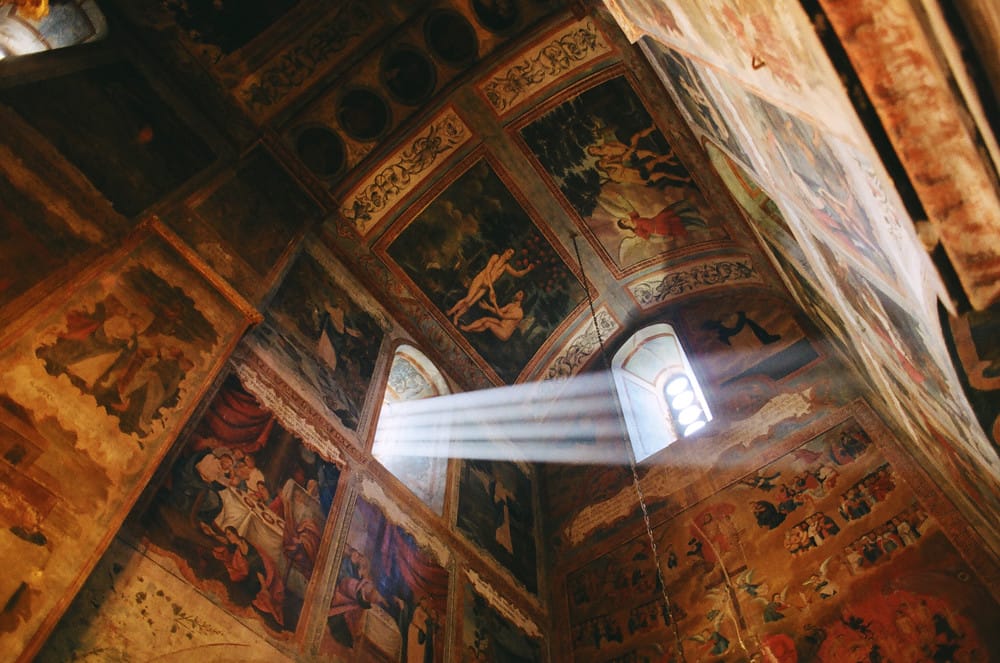 Ornate church ceiling with biblical frescoes and dramatic light beam streaming through arched window.